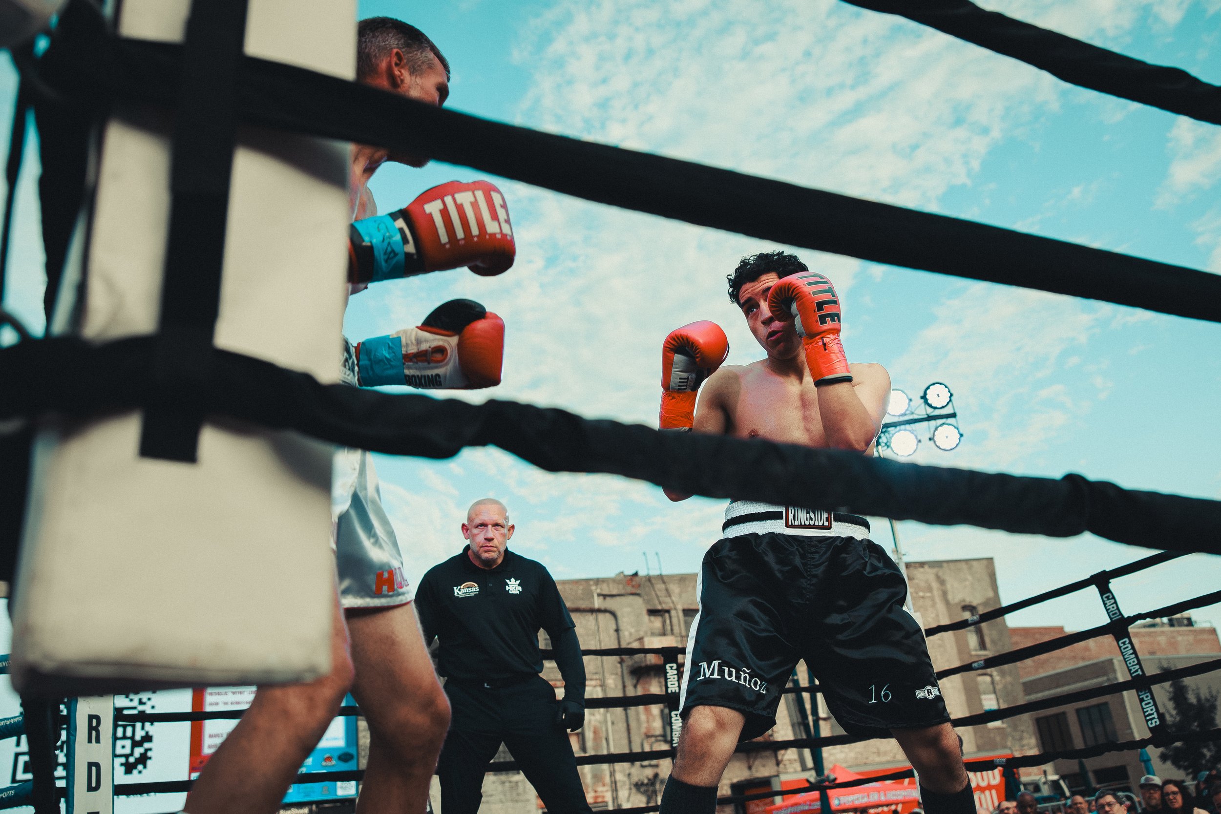 Boxers sparring in an outdoor boxing ring, with a referee observing. One boxer is shirtless, wearing orange gloves and black shorts, while the other boxer’s face is not visible. The background features a blue sky with clouds, and the scene is surroun
