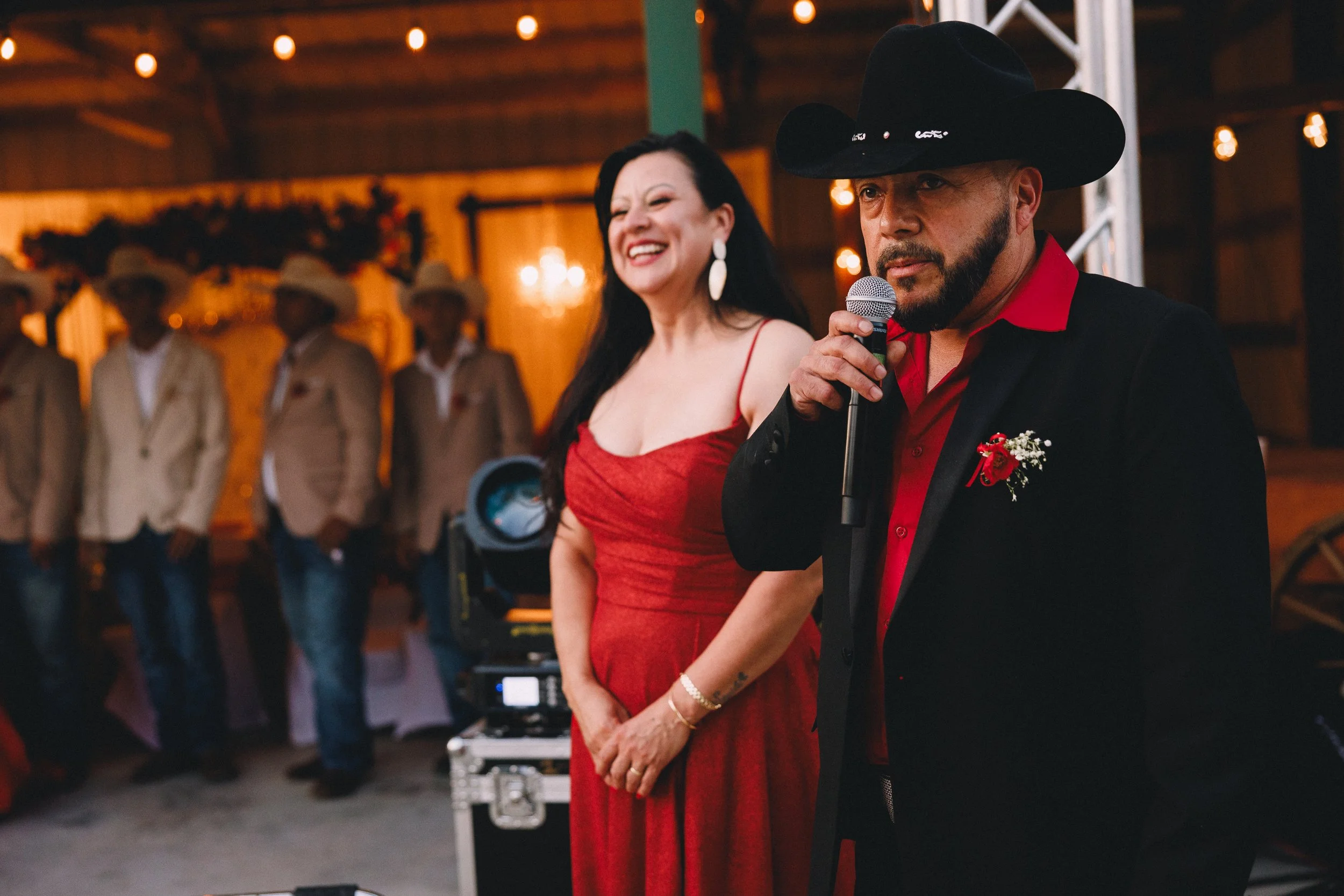 Man in cowboy hat speaking into microphone and woman in red dress smiling, at a festive event with string lights and group of people in cowboy hats in the background.