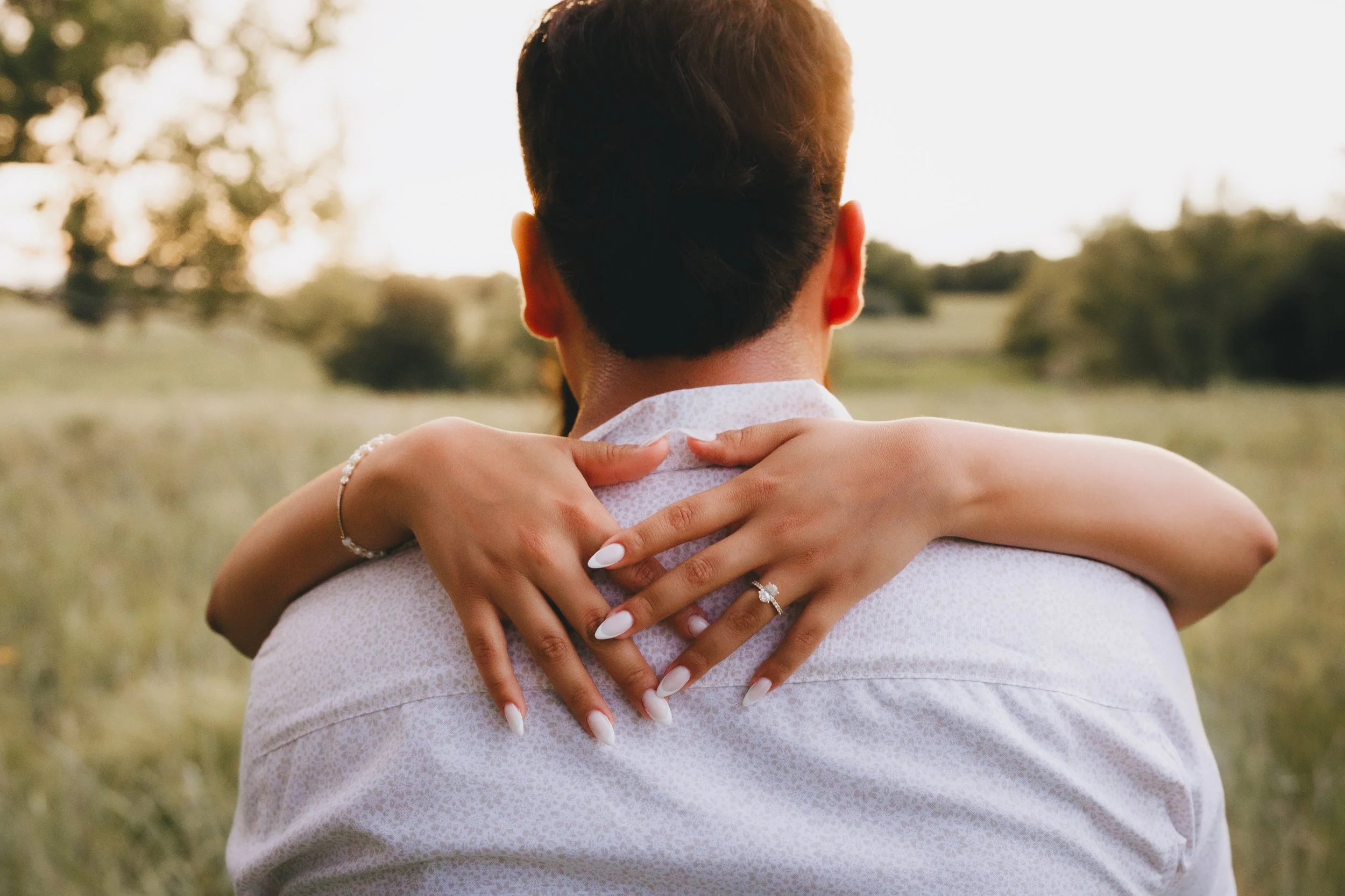 Couple embracing outdoors, the woman’s arms around the man's neck, focus on their hands and rings, with field and trees in the background.  Shot in Topeka, Kansas. 