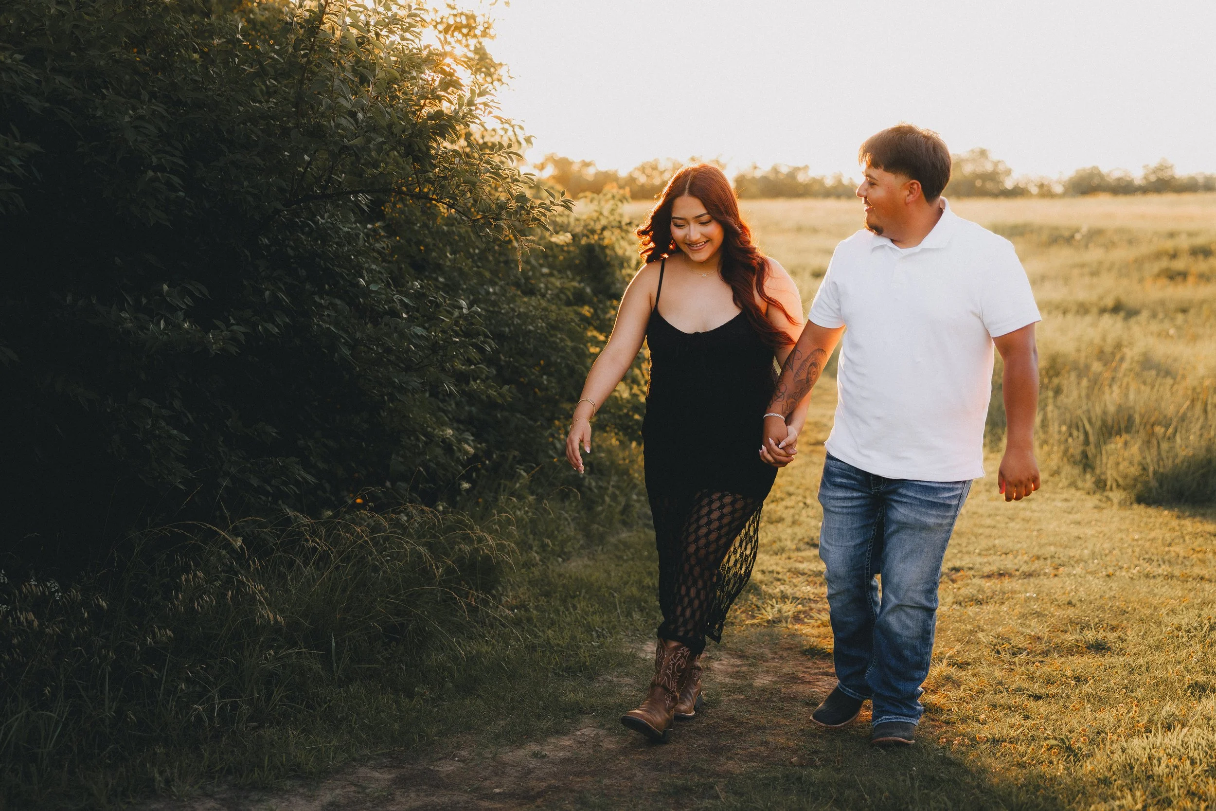 A couple walking hand in hand on a dirt path through a field at sunset, smiling and looking at each other.  Shot in Topeka, Kansas. 