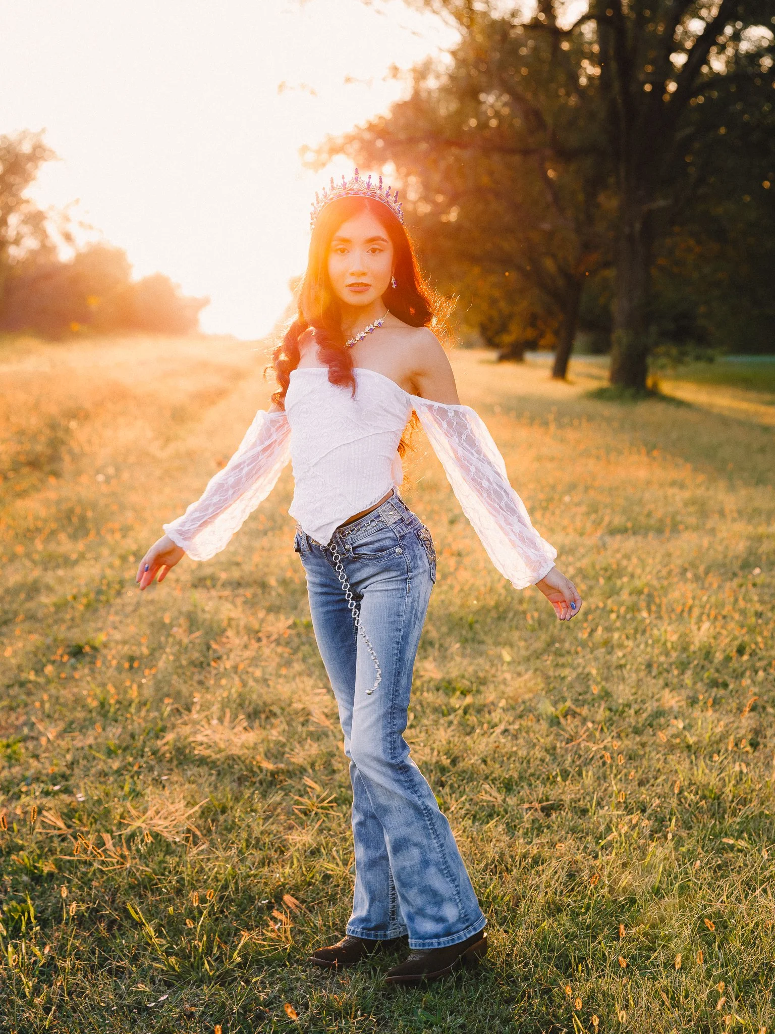 A young woman standing in a grassy field during sunset, wearing a white off-the-shoulder blouse, blue jeans, black boots, a crown, and jewelry.