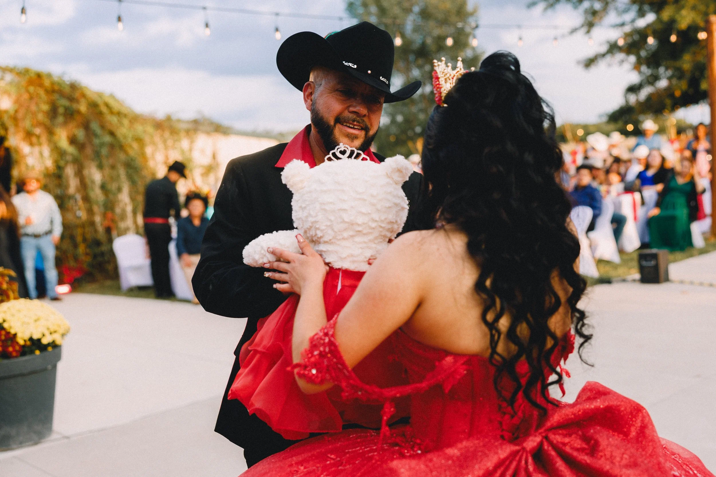 A man dressed in a cowboy hat and black suit with red shirt presents a teddy bear in a red dress to a woman in a red dress at an outdoor event with people and string lights in the background.