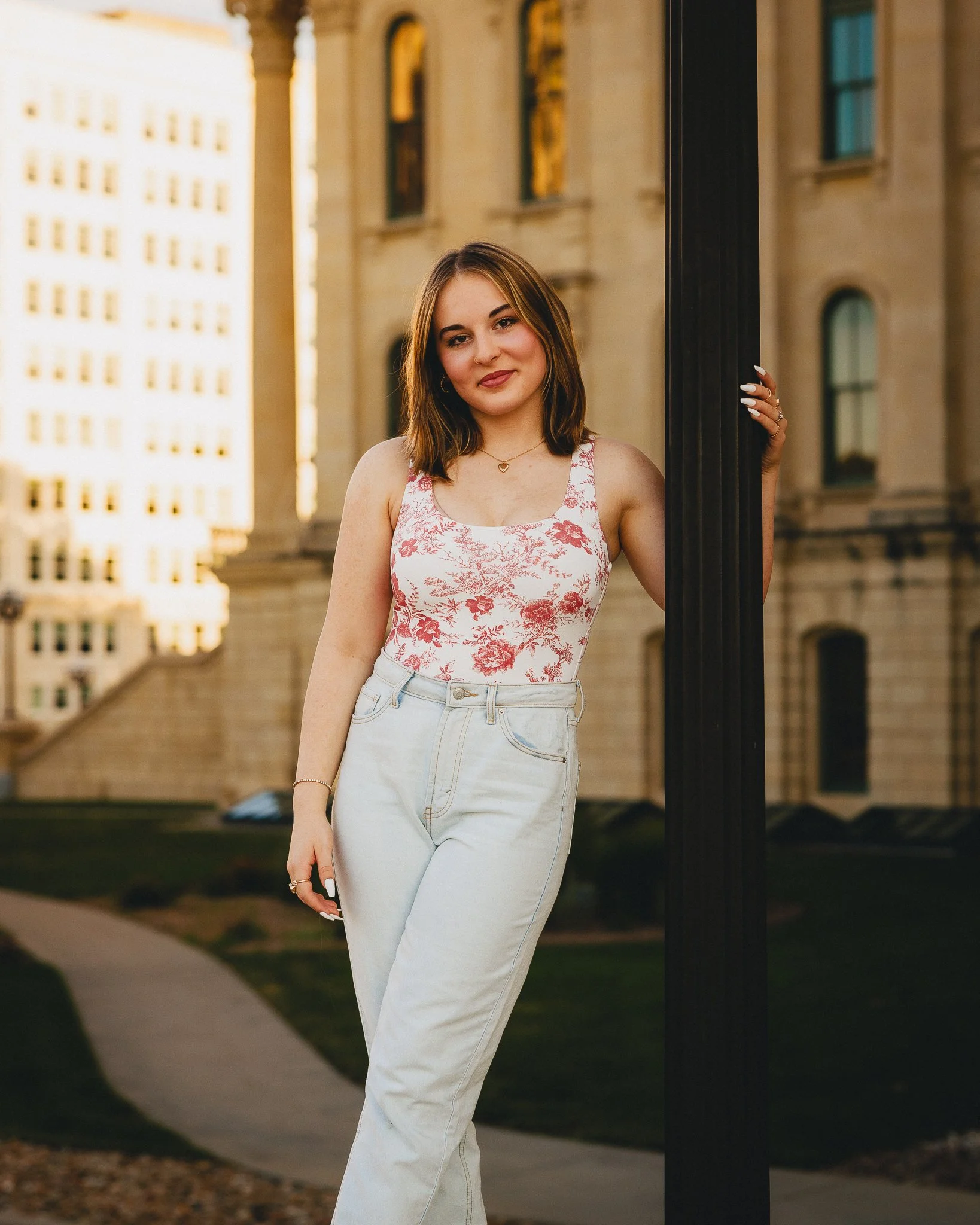 A young woman with shoulder-length brown hair, wearing a white sleeveless top with red floral pattern and high-waisted light-wash jeans, standing outdoors near a black pole with a cityscape background of tall buildings and a walking path. Shot in Top