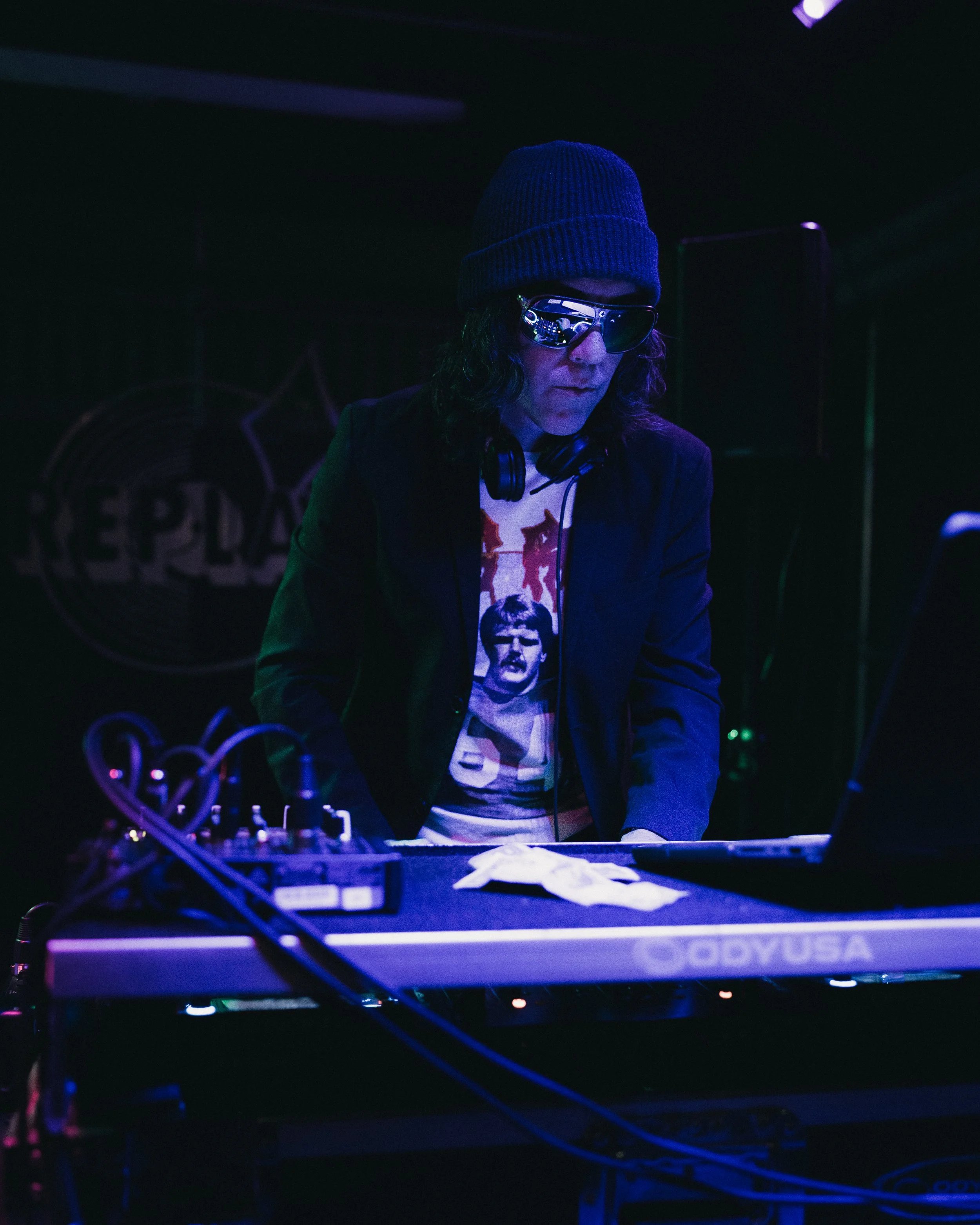 A DJ wearing sunglasses, a beanie, and headphones around his neck, DJing at a nightclub with a dark background and colorful lighting. Shot at the Replay Lounge in Lawrence, Kansas. 