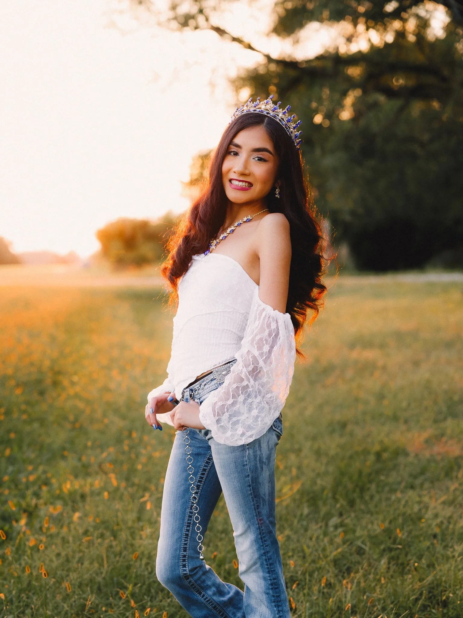 Young woman wearing a tiara, white off-shoulder blouse with lace sleeves, jeans with chain, standing outdoors during sunset, smiling.