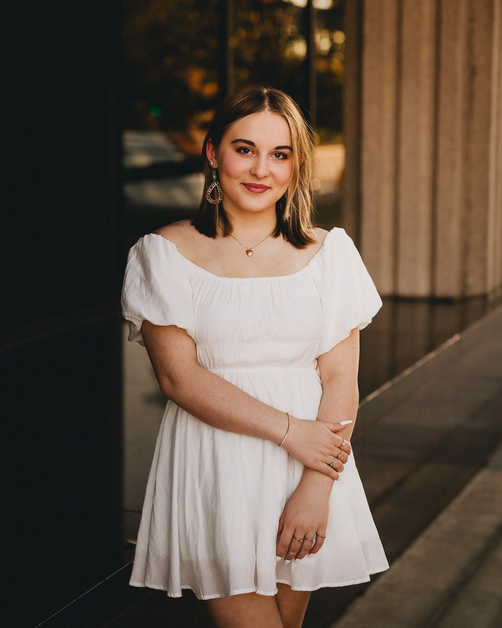 A young woman with shoulder-length brown hair wearing a white dress and colorful earrings standing outside during sunset. Shot in Topeka, Kansas. 