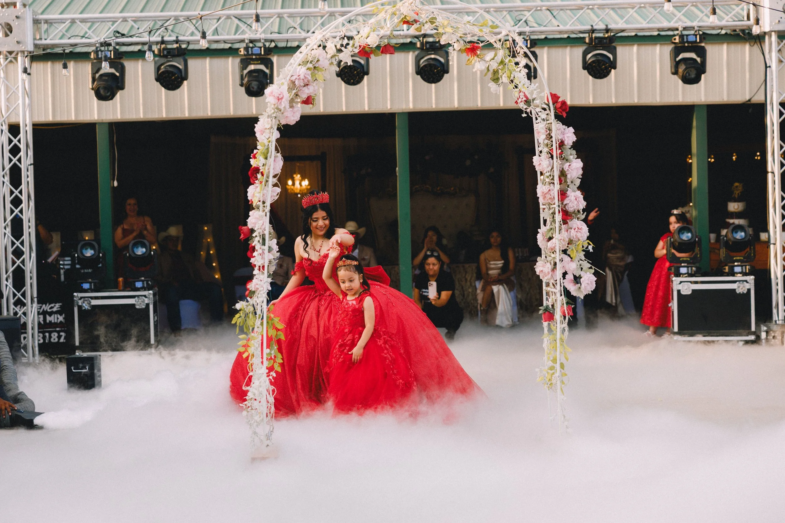 A woman and a young girl in red dresses dancing under a floral arch at a wedding reception with fog effects and guests watching in the background.