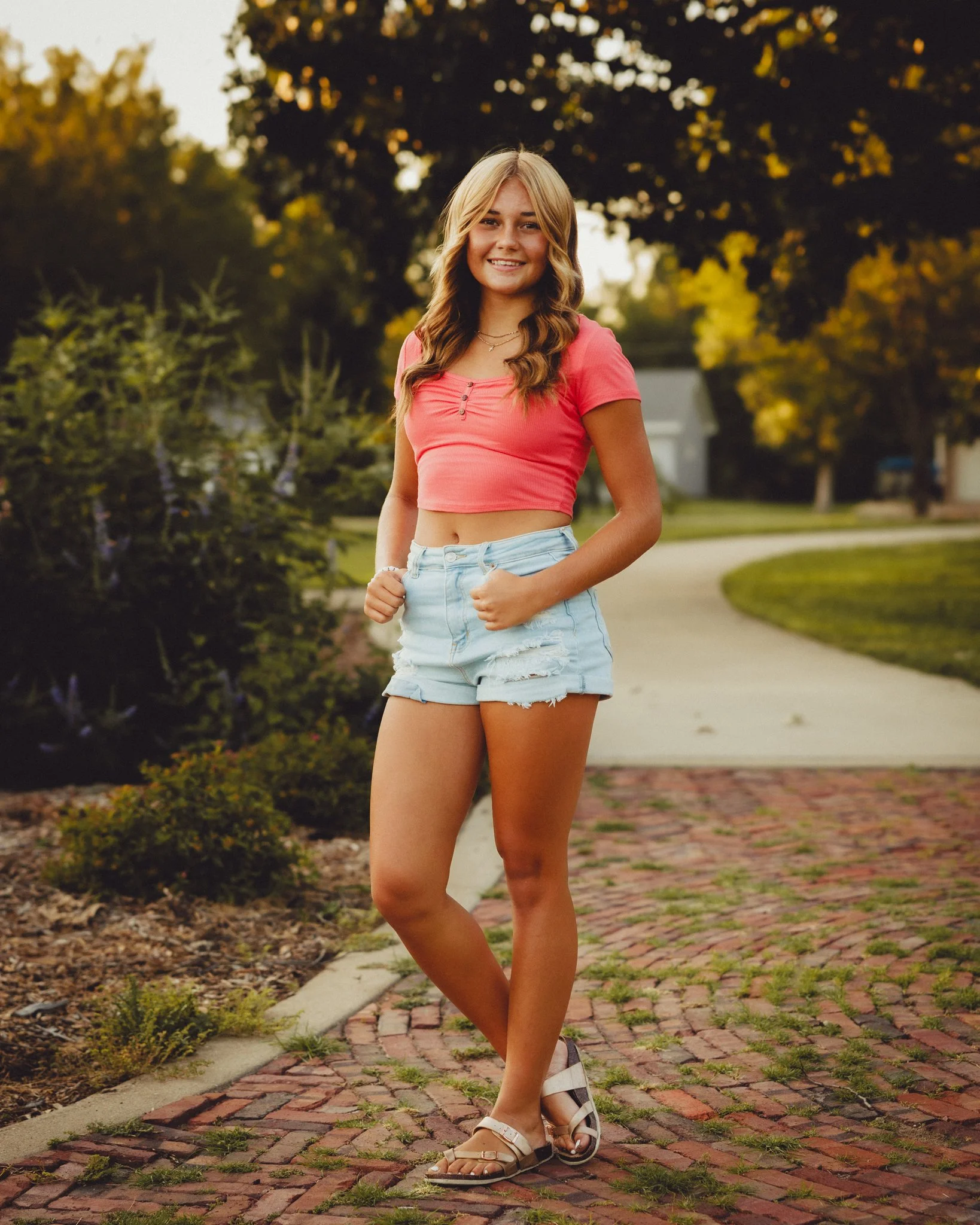 A young woman in a pink crop top and ripped denim shorts standing outdoors on a brick sidewalk, smiling at the camera, with trees and houses in the background during sunset. Shot in Topeka, Kansas. 
