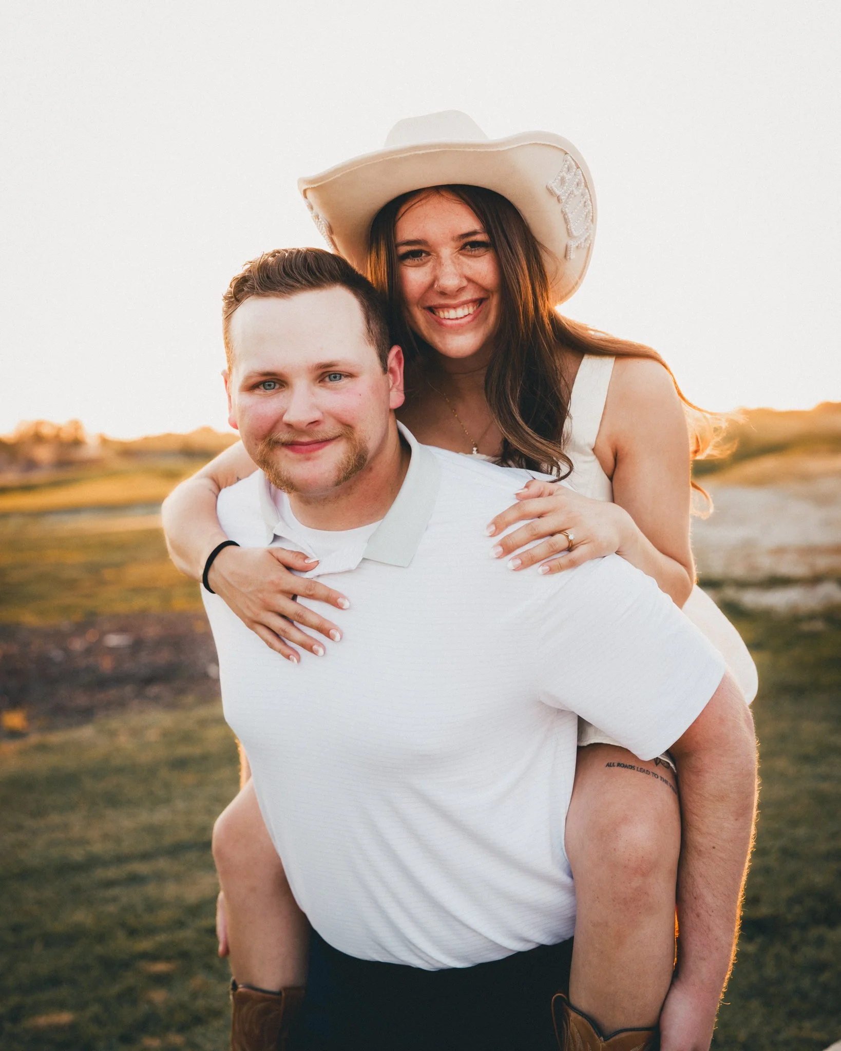 A smiling couple outdoors during sunset, the woman is on the man's back, wearing a cowboy hat and a white dress, with her arm around his shoulders. The man is in a white polo shirt, with a small tattoo visible on his forearm.