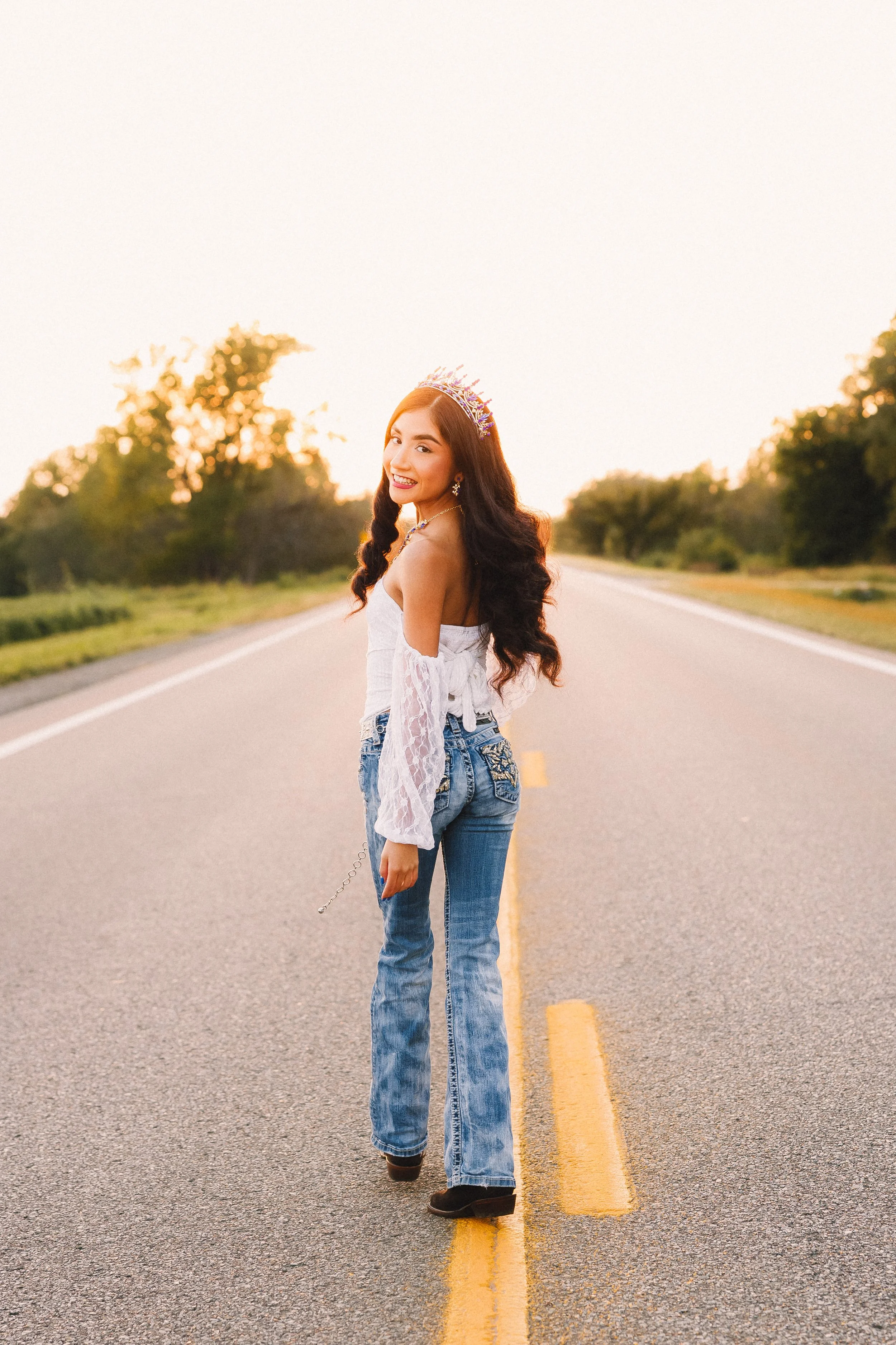 A young woman standing in the middle of an empty country road at sunset, wearing a tiara, white off-the-shoulder blouse, and blue jeans, smiling at the camera.