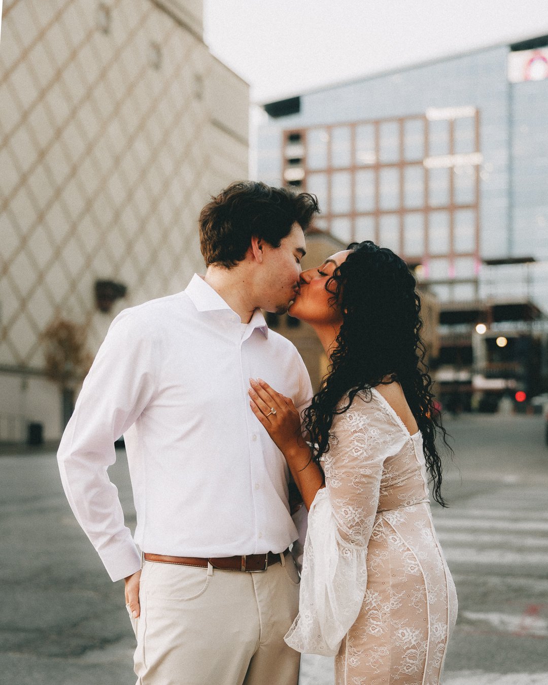 A couple kissing outdoors in an urban area, with the woman wearing a sheer lace dress and the man dressed in a white shirt and beige pants. Shot at Kansas City Country Club Plaza. 
