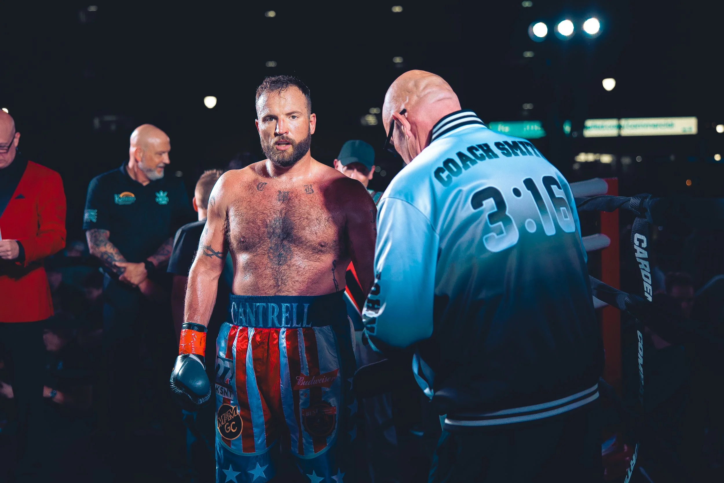 A shirtless male boxer wearing American flag-themed shorts and boxing gloves standing inside a boxing ring, talking to a referee wearing a jacket with the words Coach Smith and the time 3:16 on the back, while other officials and spectators look on i