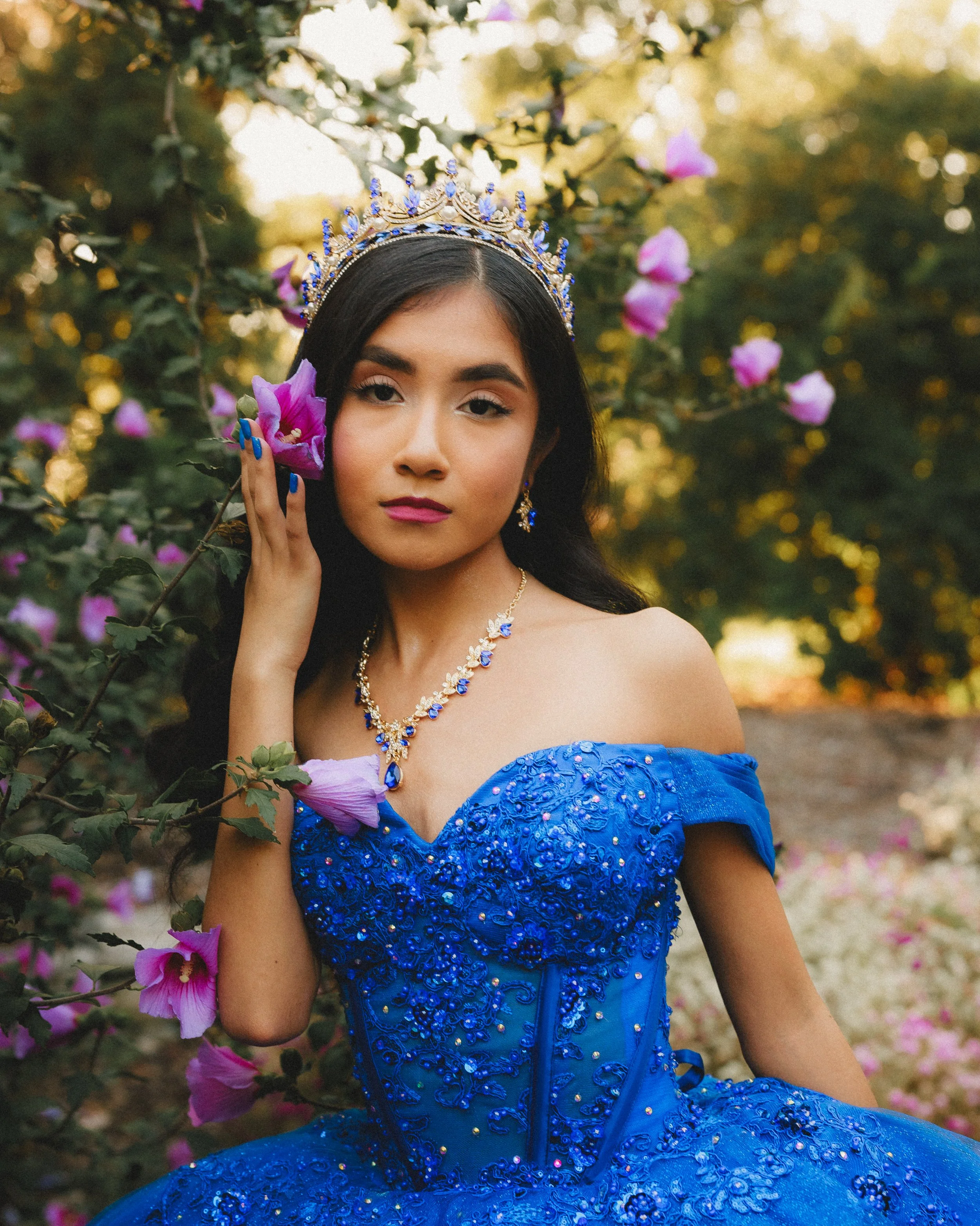Young woman wearing a regal blue dress with intricate beadwork, a tiara, and matching jewelry, standing amidst pink and purple flowers outdoors.