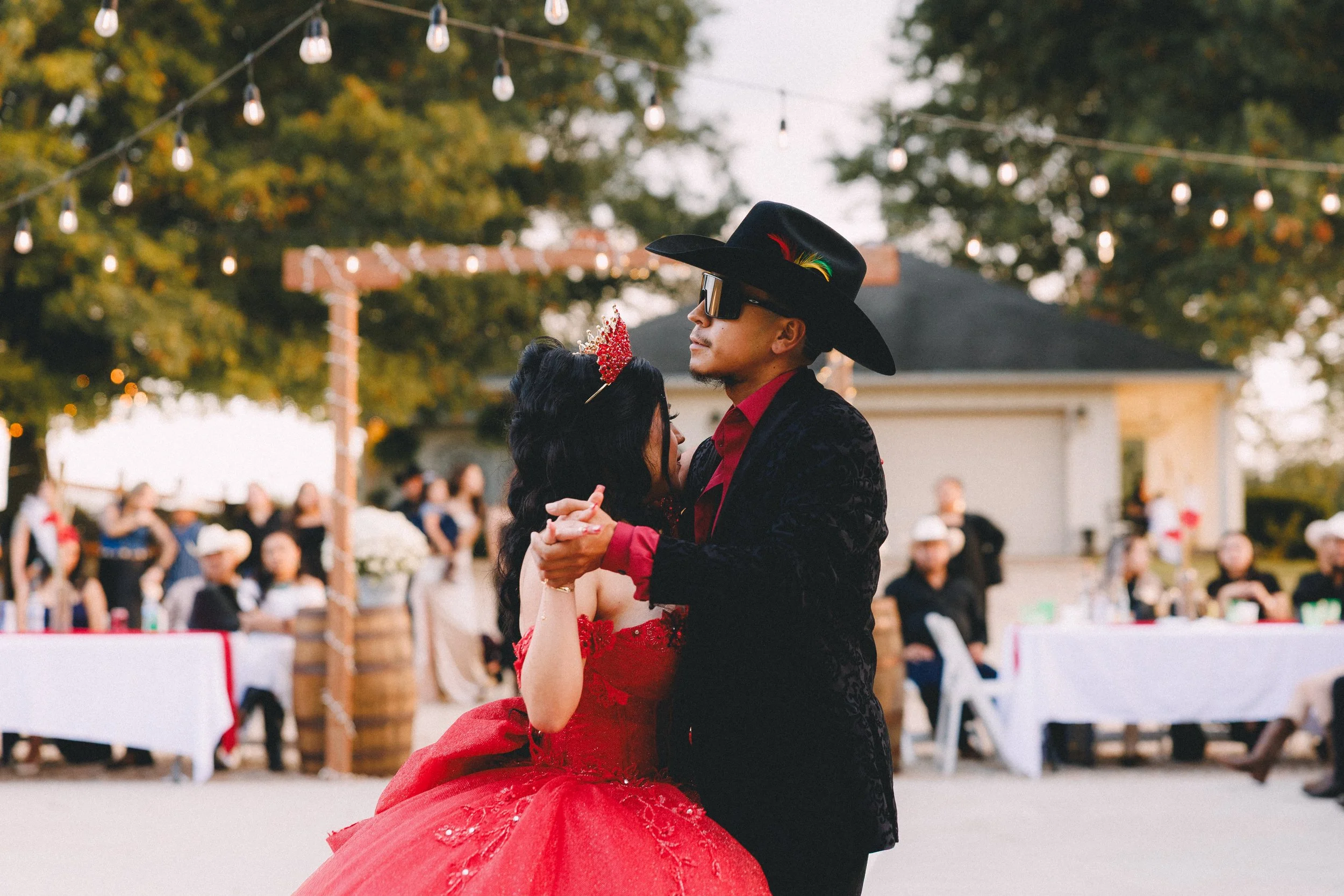 A couple is dancing outdoors at a celebration or party, with the woman in a red gown and the man in a black jacket, red shirt, large black hat with colorful feathers, and sunglasses.