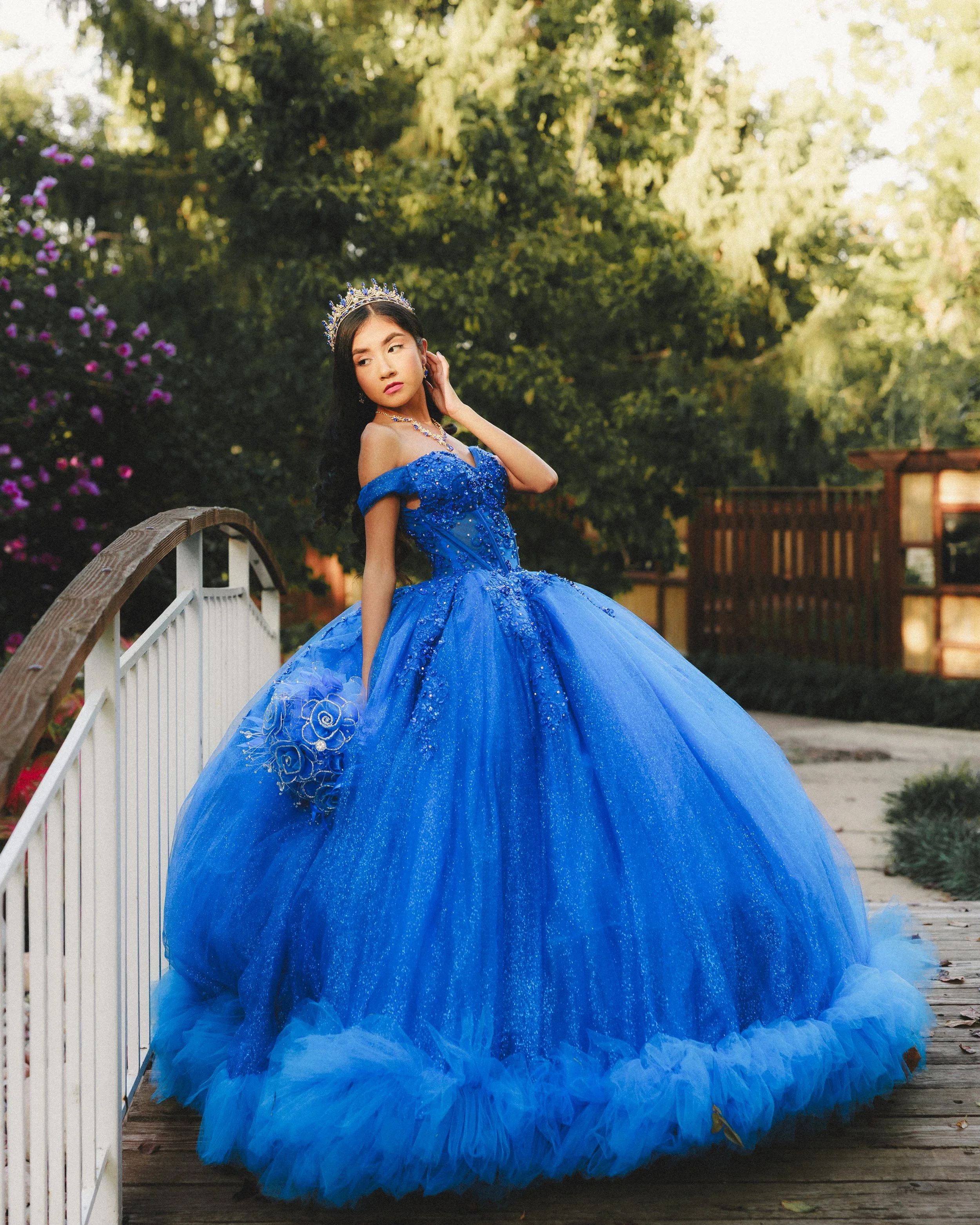 Young woman in a sparkling blue princess gown standing on a wooden bridge in a garden with greenery and flowers, wearing a tiara and jewelry.
