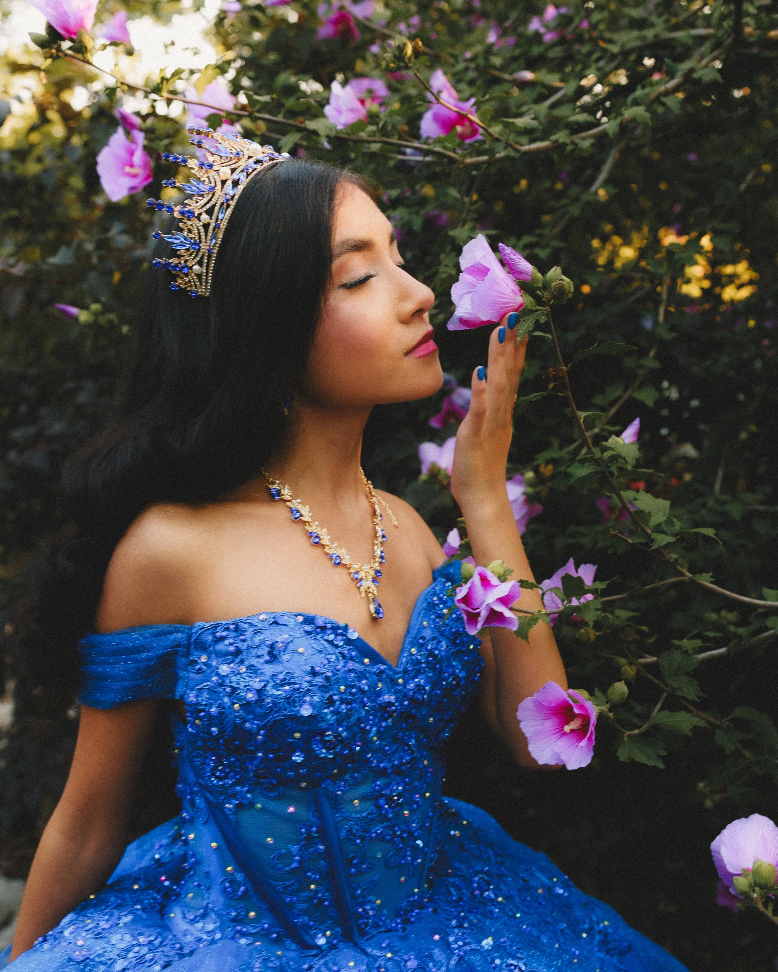 A young woman dressed in a royal blue, beaded, off-shoulder gown with intricate embroidery and embellishments, sitting among pink and purple flowers, gently holding a flower close to her face with her eyes closed, wearing a matching blue and gold tia