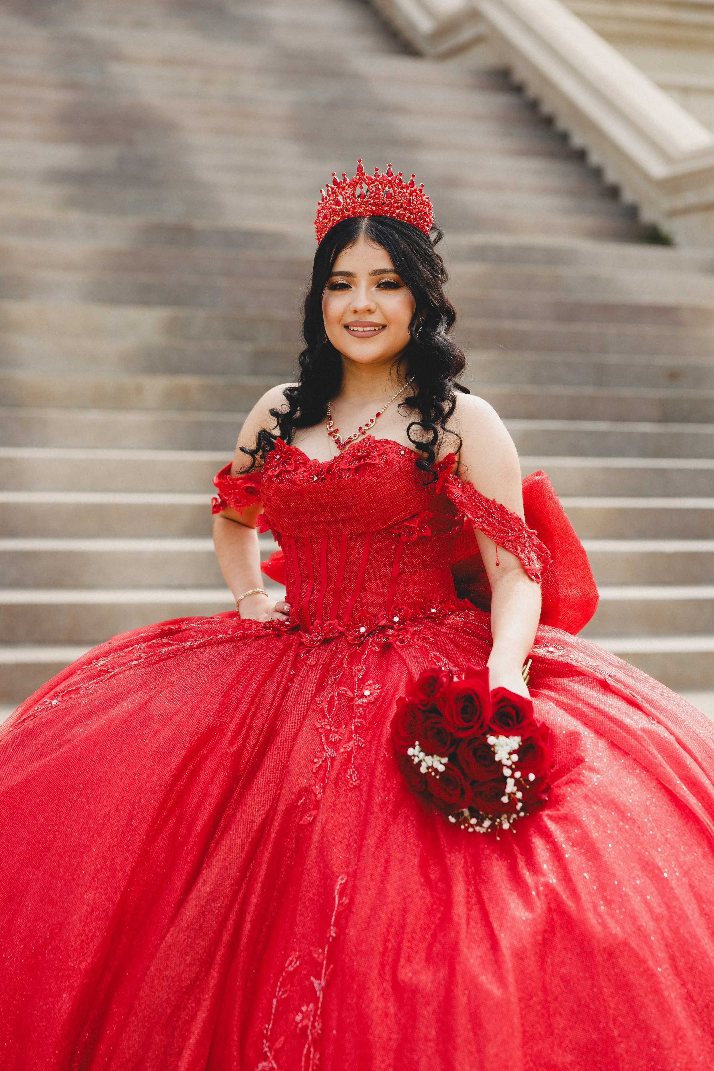 A woman in an elaborate red gown with lace and embroidery details, wearing a red crown and holding a bouquet of red roses, standing on outdoor steps.
