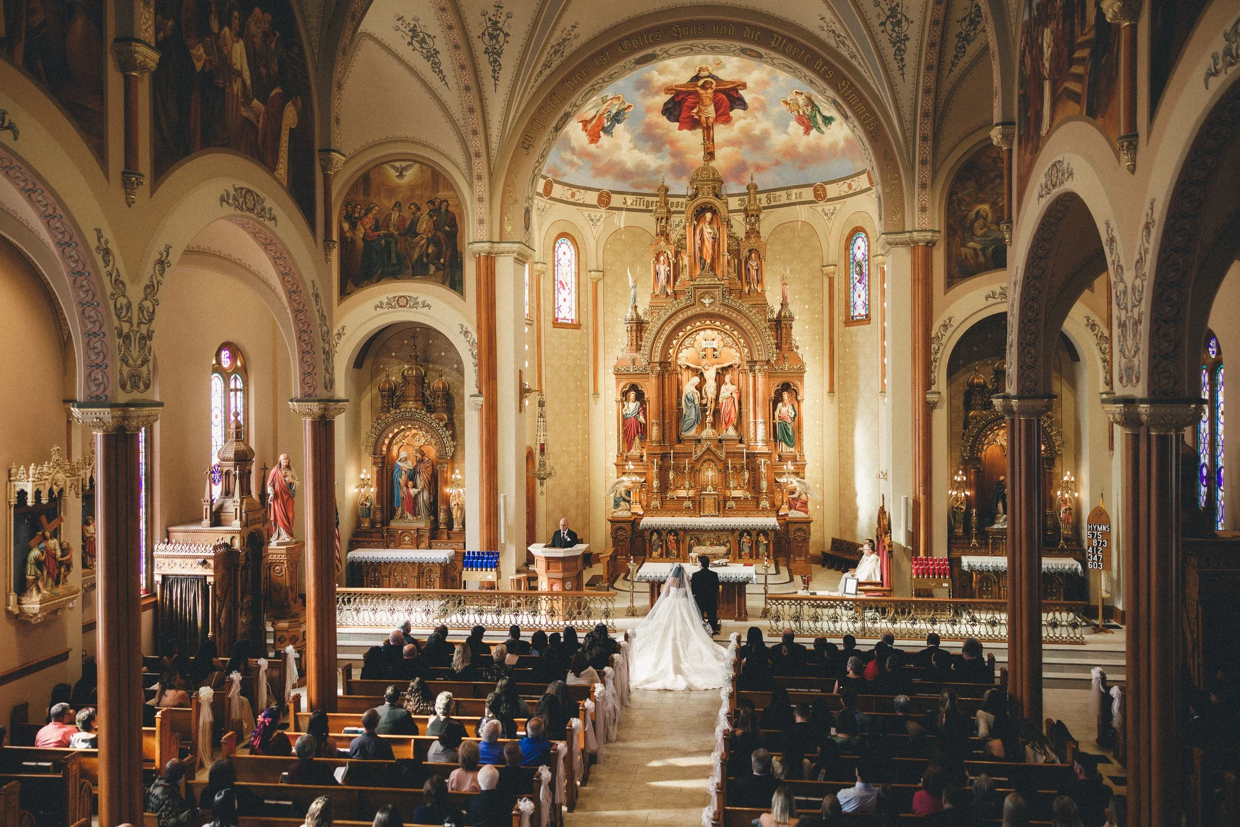 A wedding ceremony in a church with a bride and groom standing at the altar. The church interior features ornate decorations, religious statues, stained glass windows, and a painted ceiling. Guests are seated in pews, watching the ceremony.