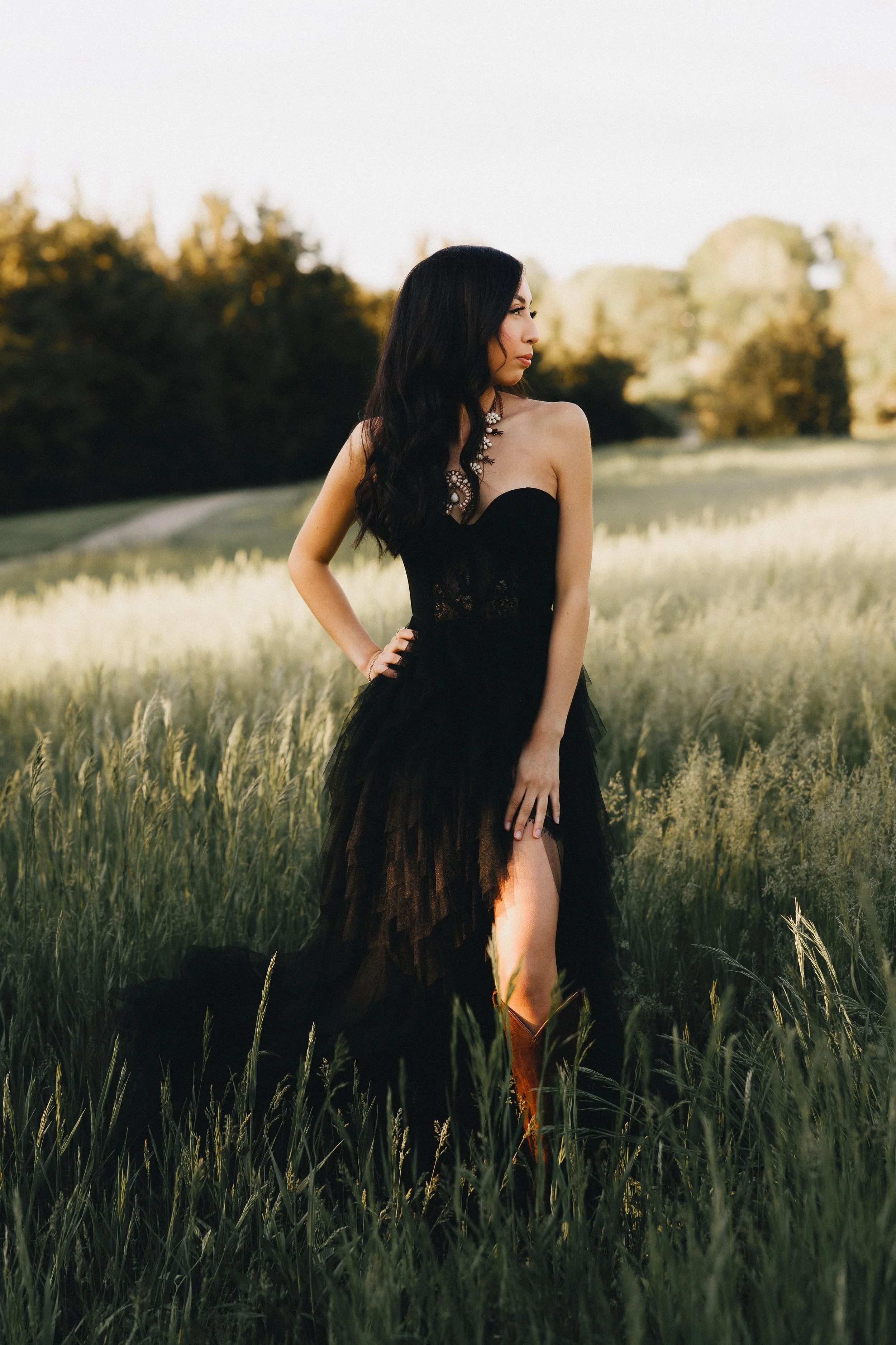 A woman with long black hair wearing a strapless black dress with a high slit, standing in a grassy field during sunset.