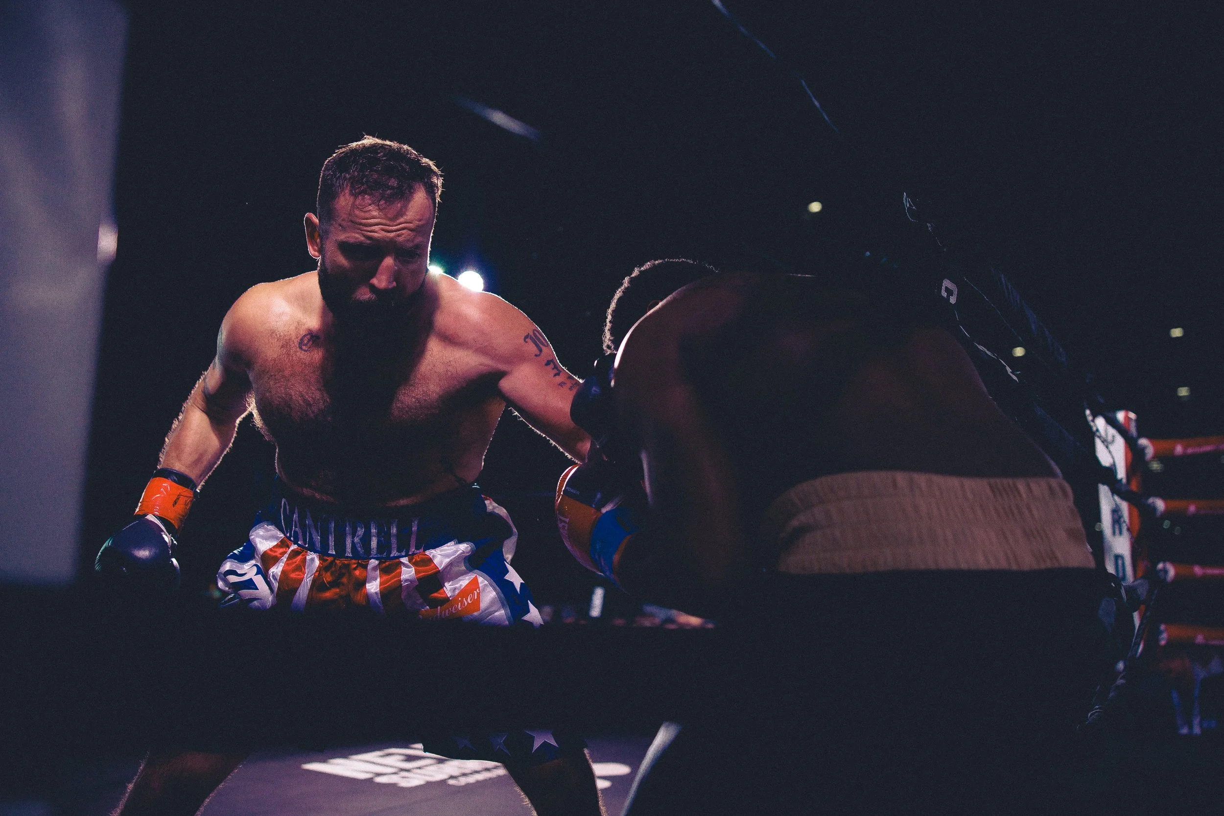 Two male boxers in a boxing ring during a match, one is leaning forward and delivering a punch to the other, who is bent over in defense. It is dark with bright lights overhead. Shot at Evergy Plaza in Topeka, Kansas. 