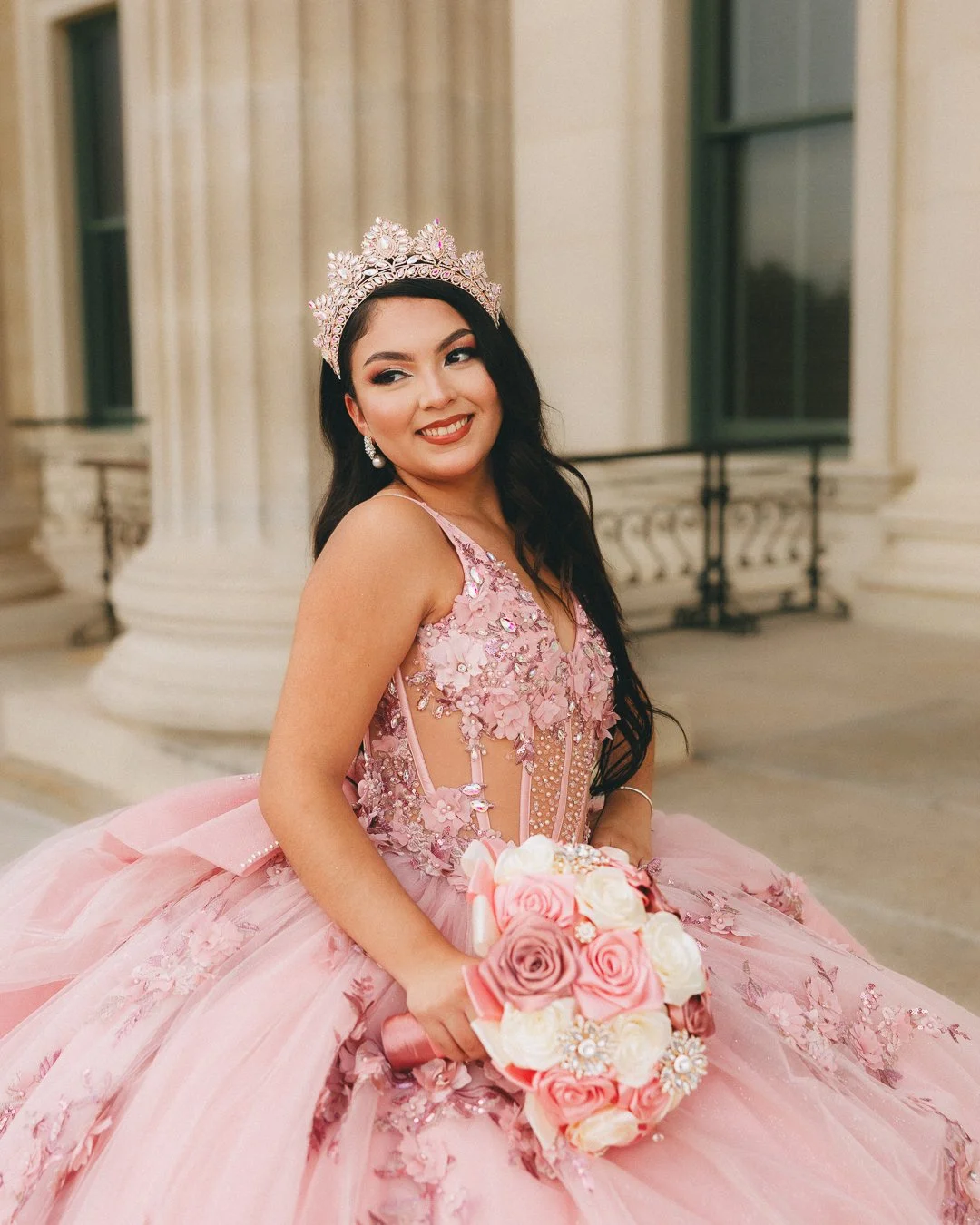 A woman in a pink quinceañera dress with floral embellishments and a tiara, holding a bouquet of roses, smiling indoors near columns.