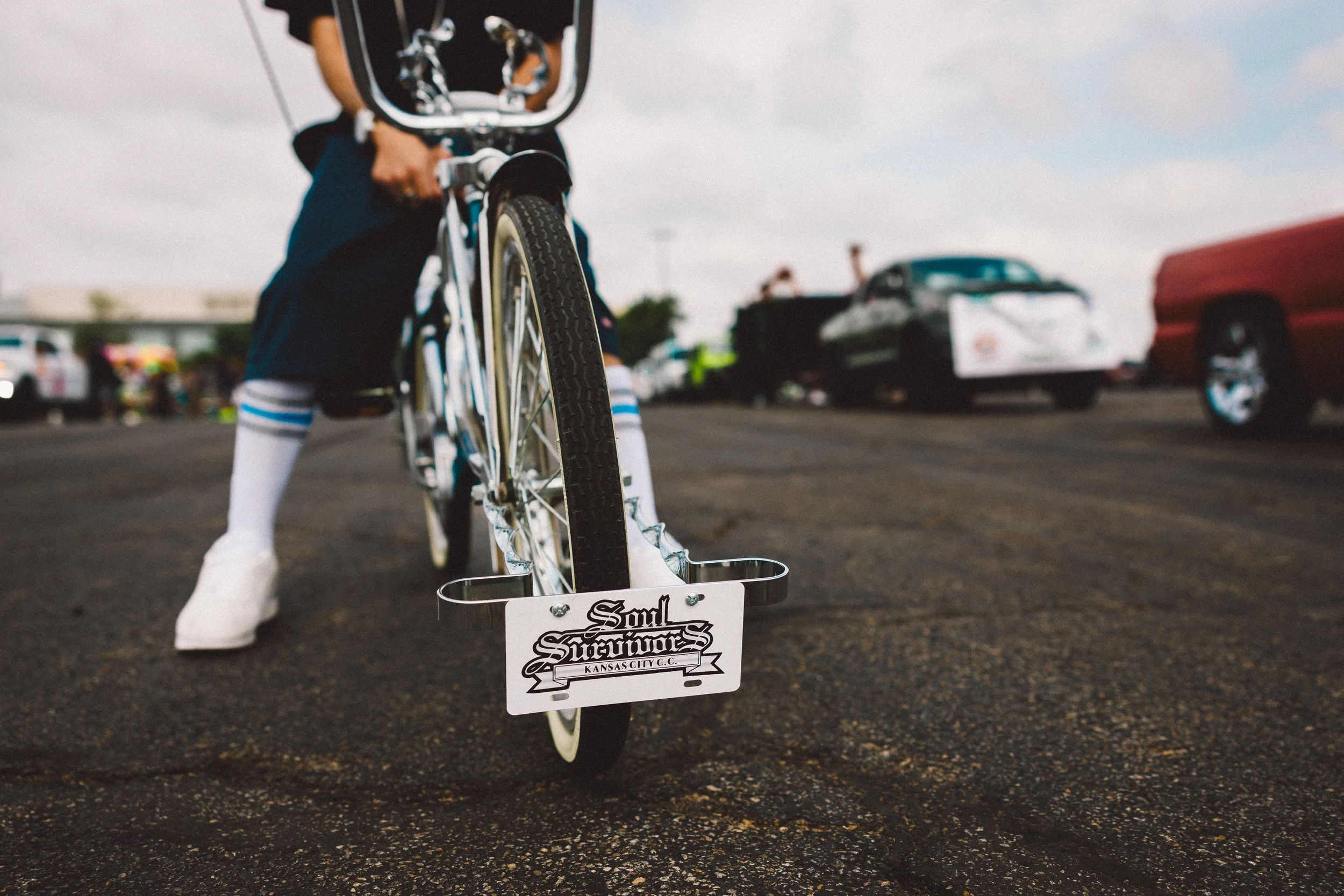 Close-up of a person riding a bicycle, focusing on the front wheel and pedal area, with several cars parked in the background. Shot at Fiesta Topeka parade.