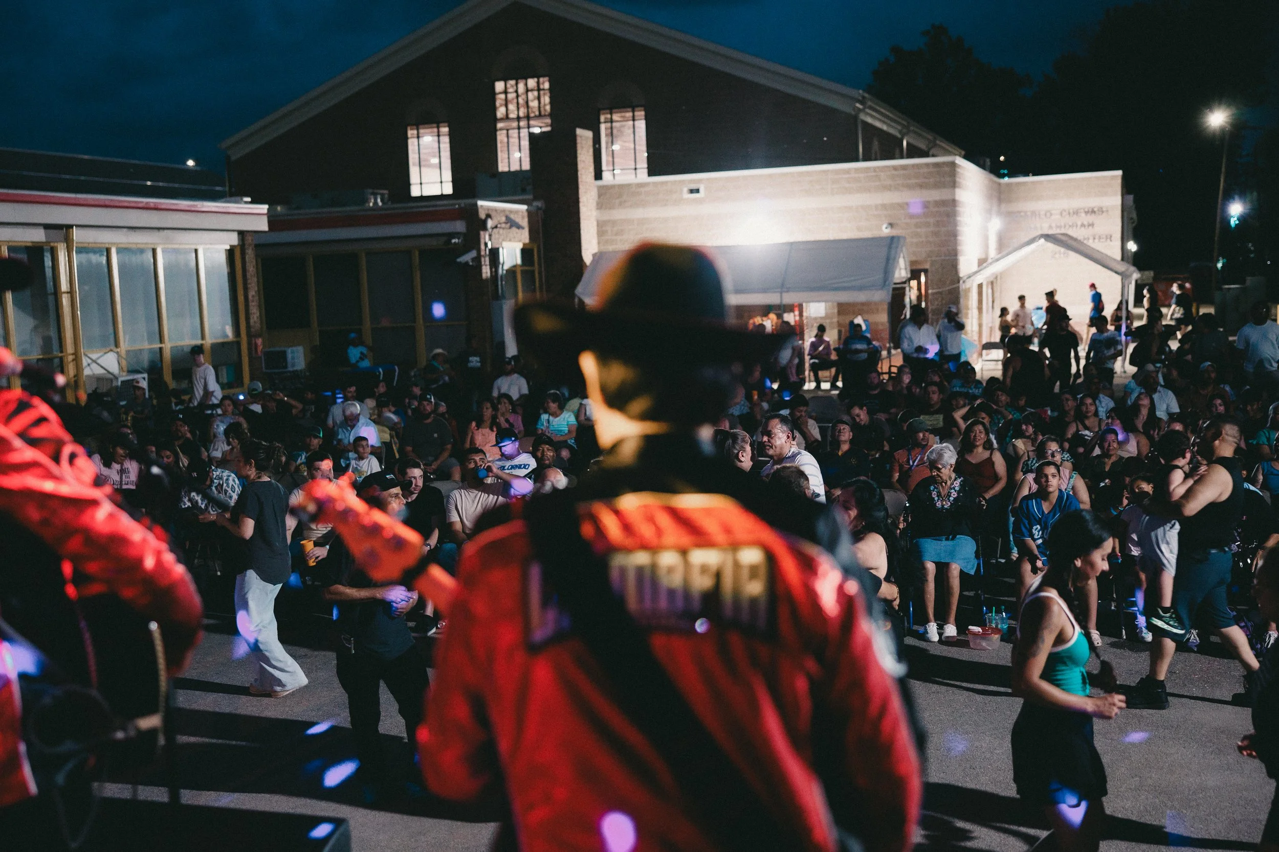 A crowd gathered outside a building during the night, with a person dressed in cowboy attire in the foreground, facing away from the camera. Shot at Fiesta Topeka.