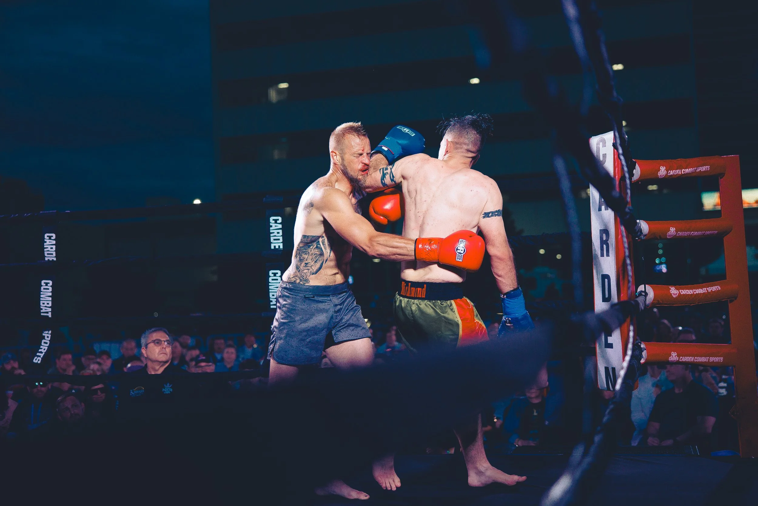 Two male fighters engaged in a boxing match inside a ring, with one delivering a punch to the other's face. The audience watches intently in the background. Shot at Evergy Plaza in Topeka, Kansas. 
