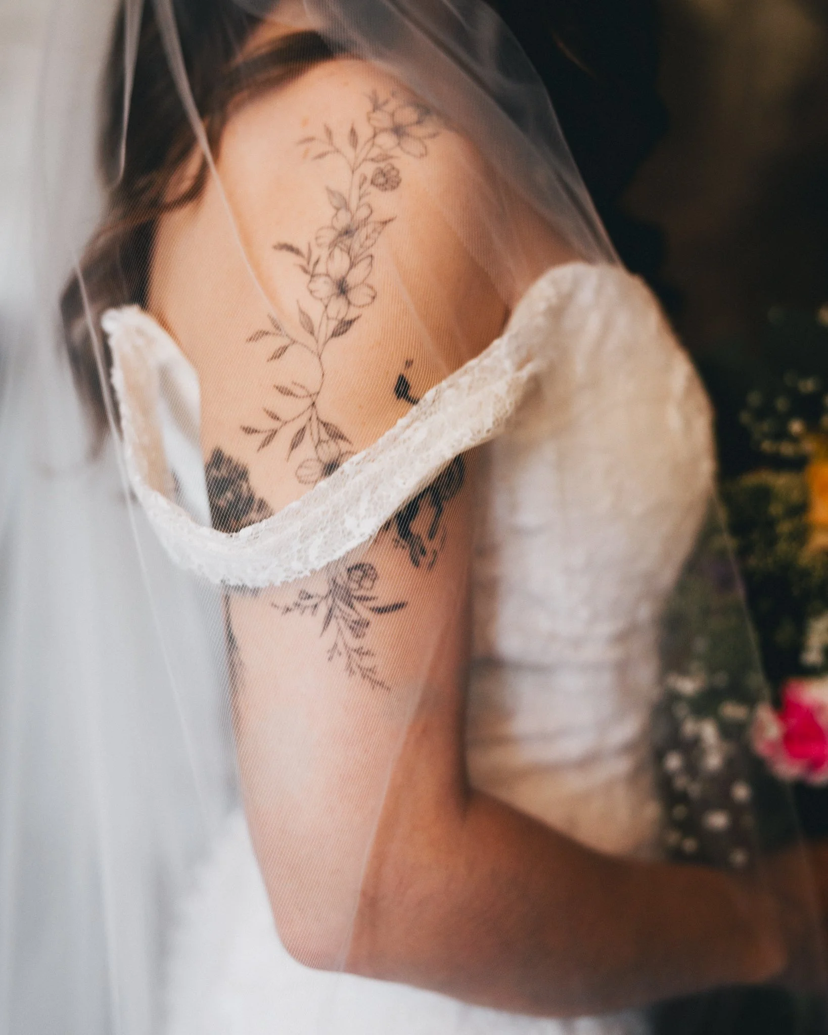 A bride wearing a lace face mask and a white lace dress, with floral tattoos on their arm and shoulder, lying down with a floral arrangement nearby.
