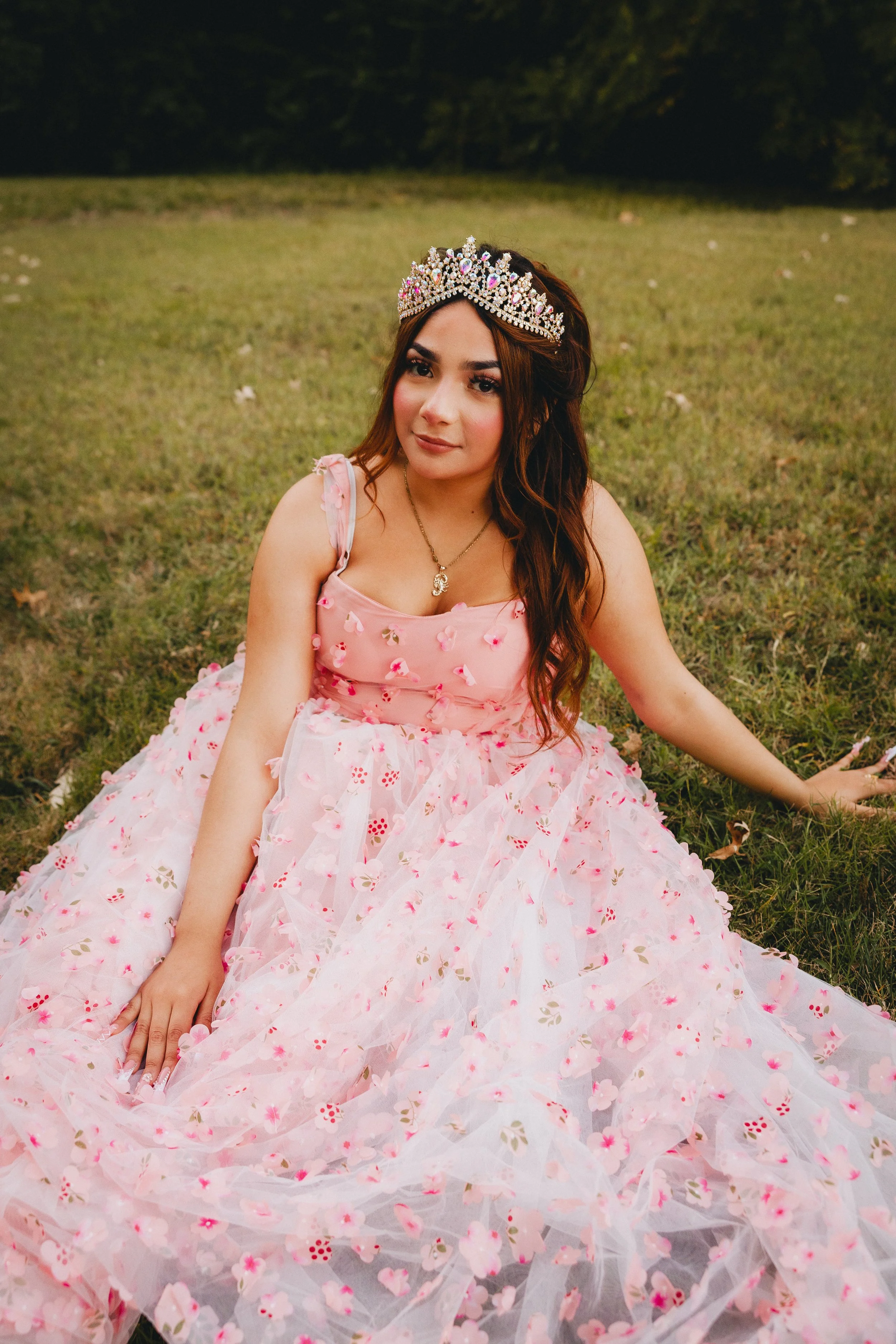 Young woman wearing a pink floral dress and a jeweled tiara, sitting on grass outdoors during daytime.
