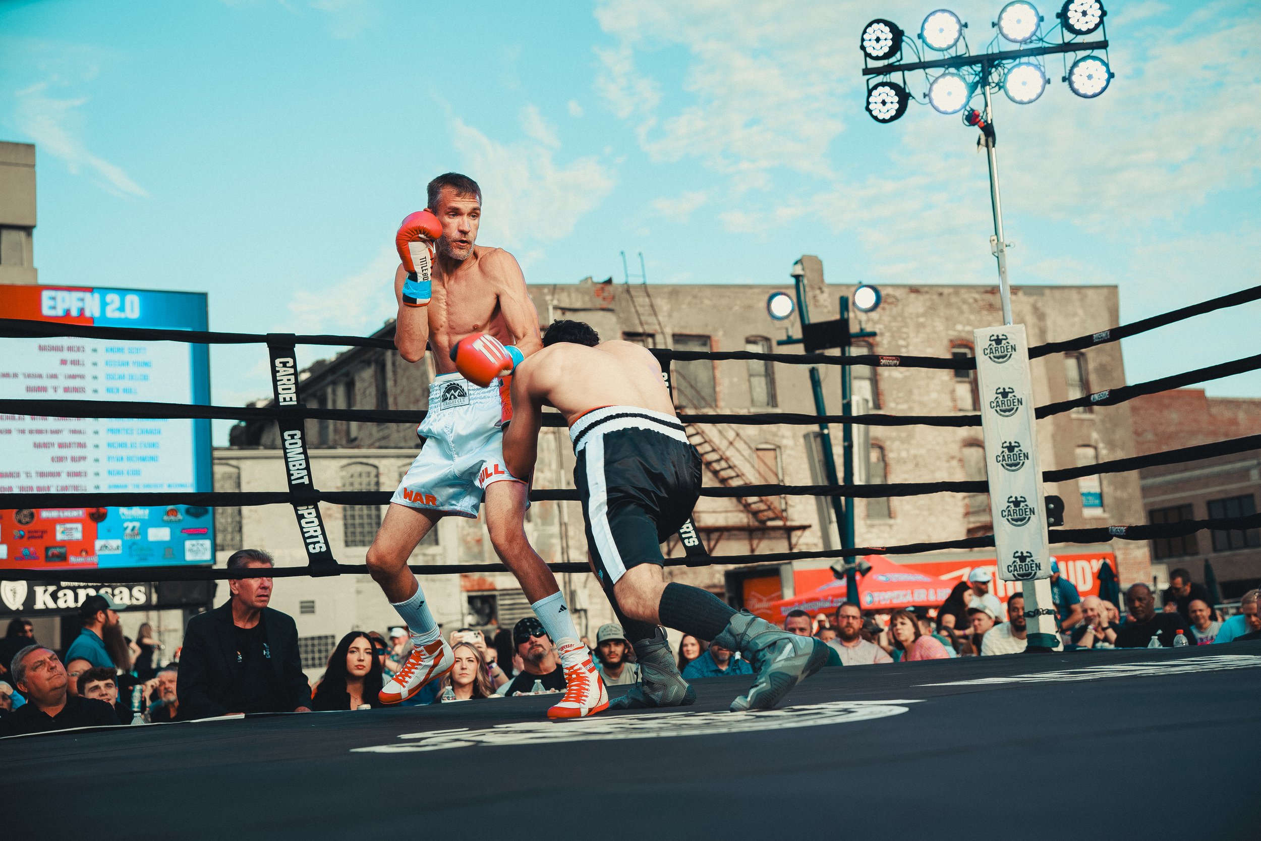 Two men boxing in an outdoor ring crowded with spectators, with buildings and a blue sky in the background. Shot at Evergy Plaza in Topeka, Kansas. 