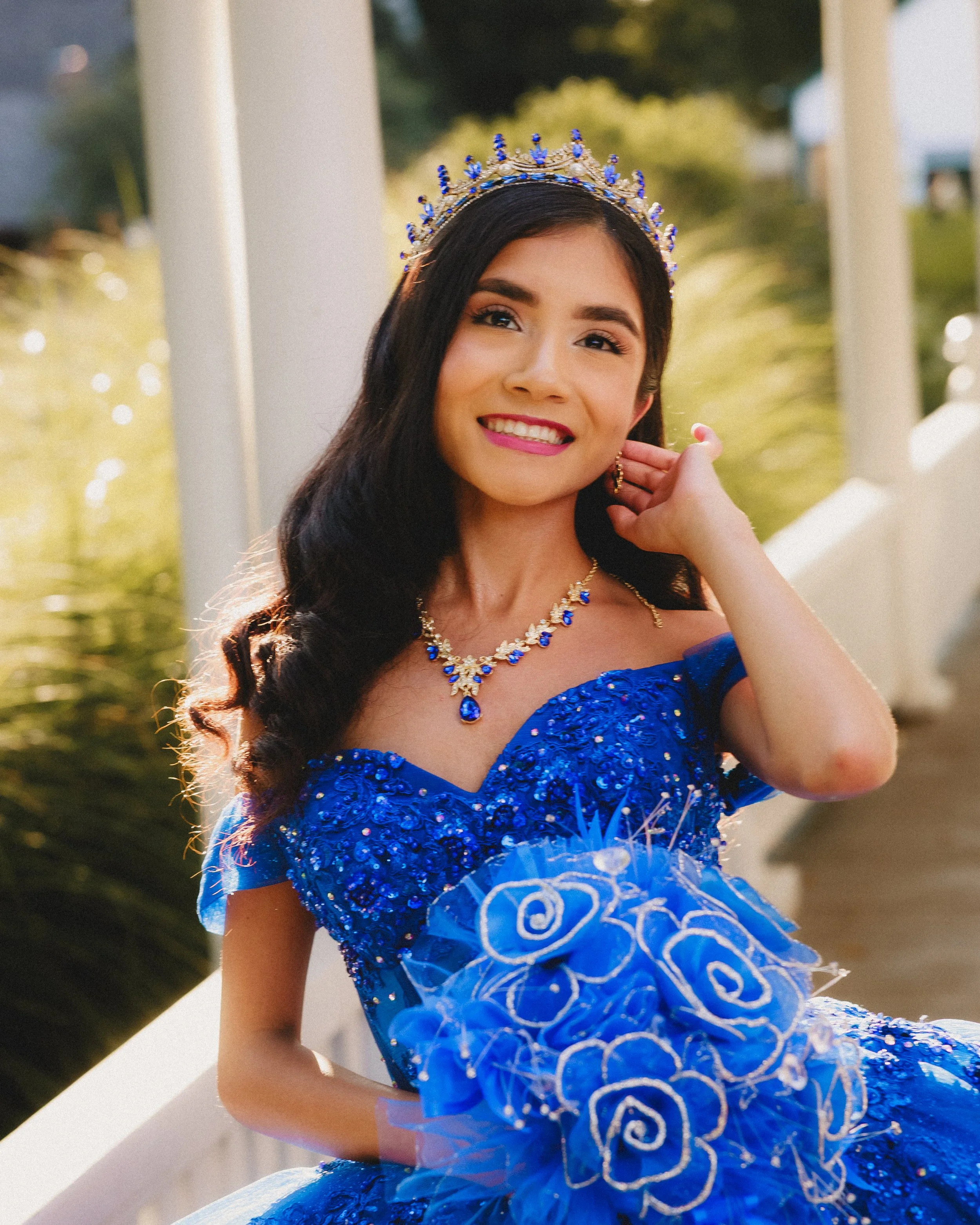 Young woman in a blue dress and jewelry, wearing a tiara, smiling outdoors.