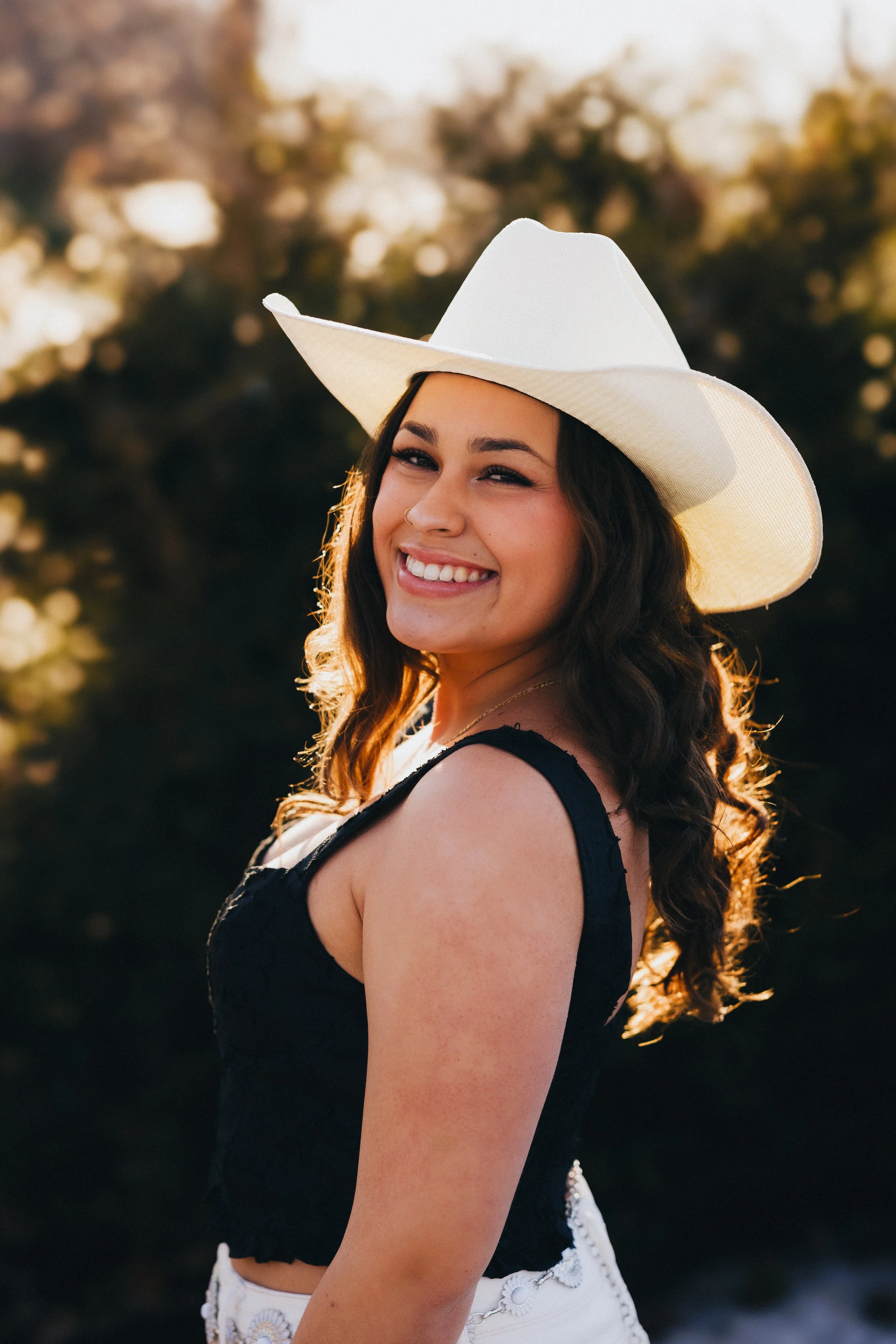A smiling woman with long dark hair in loose waves, wearing a large white sunhat and a black sleeveless top, standing outdoors in the sunlight with a blurred background of trees.