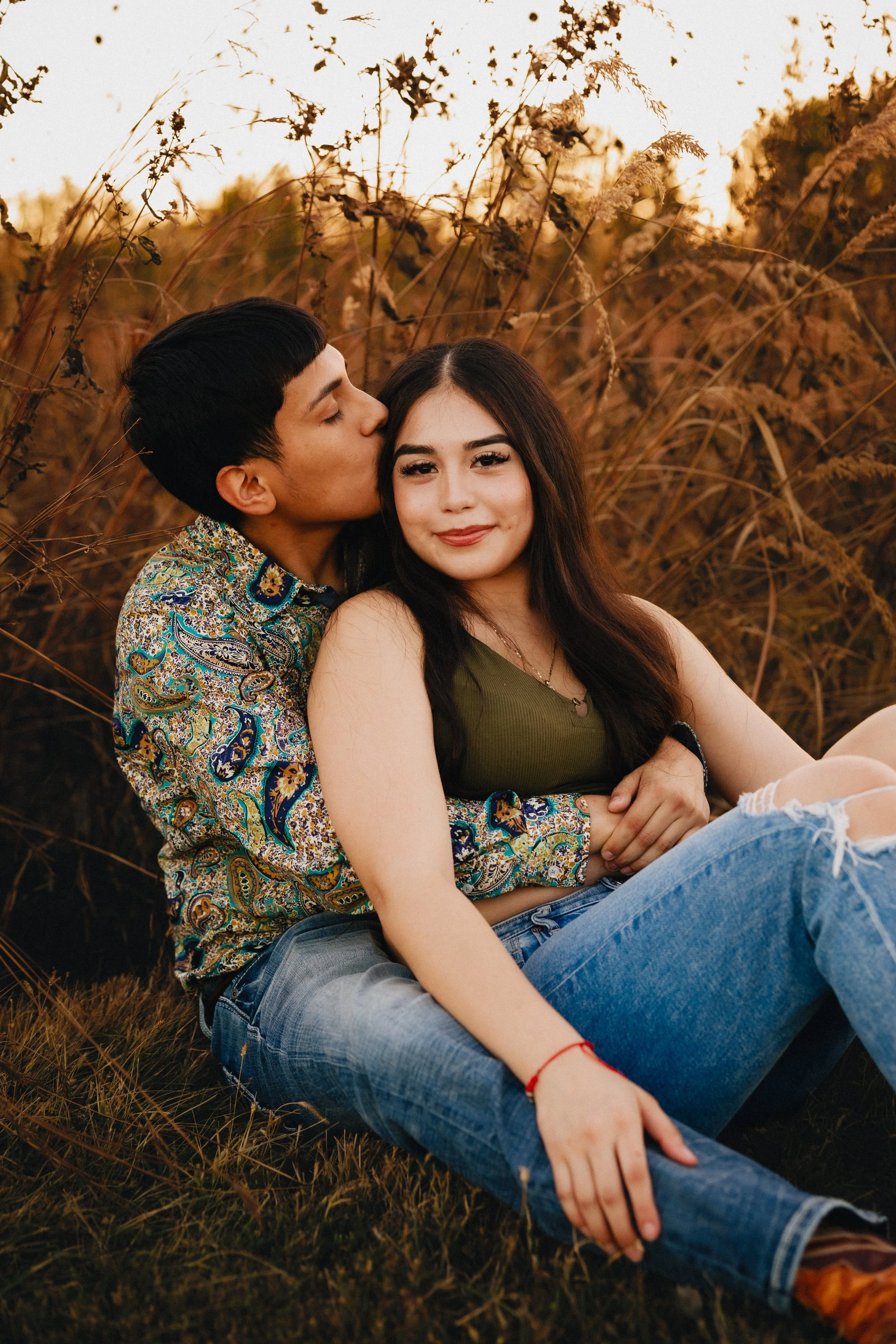 A young couple sitting outdoors in a field of tall, dry grass, with the man kissing the woman on the cheek and the woman smiling at the camera during sunset. Shot in Topeka, Kansas. 
