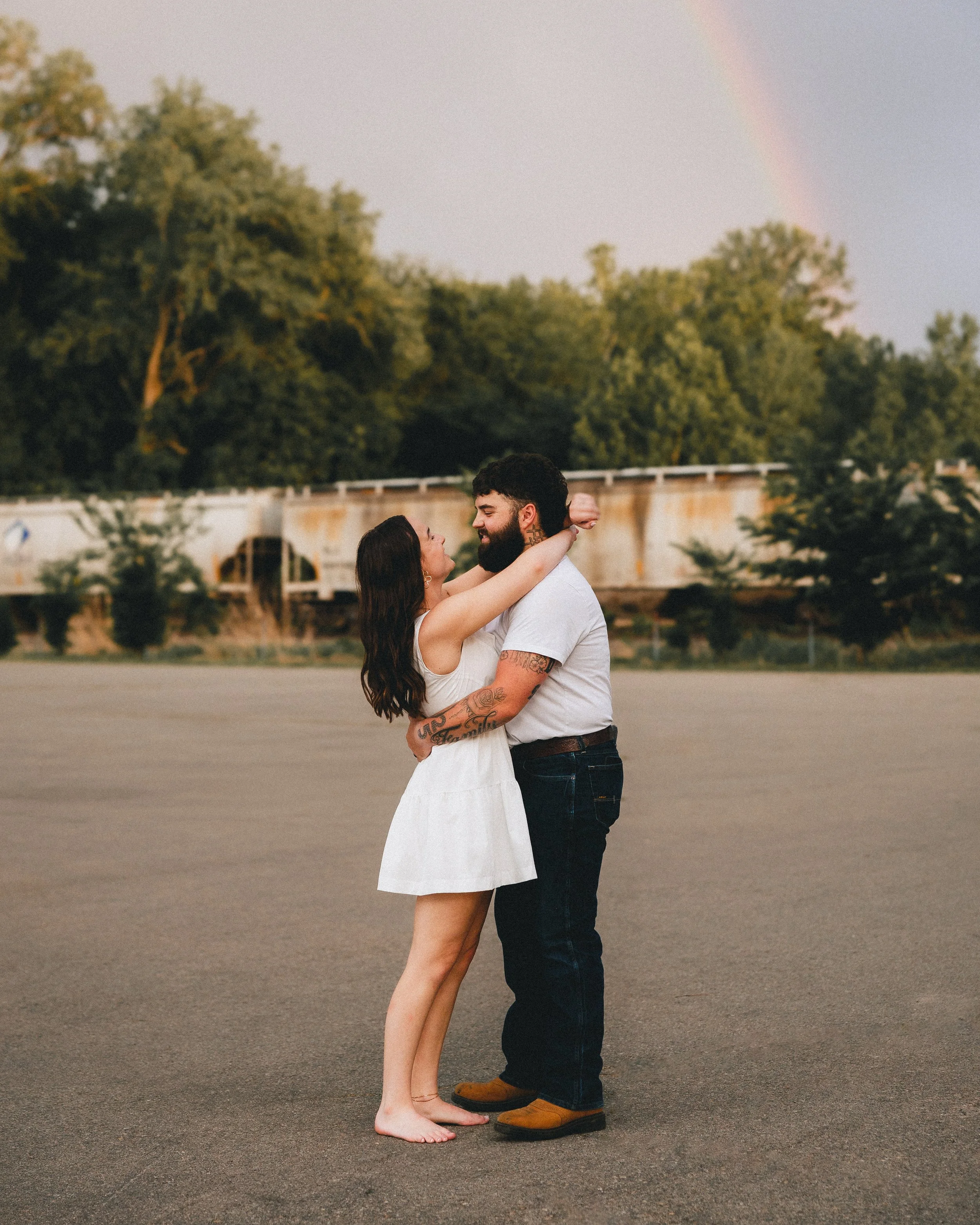 A couple embraces and looks into each other's eyes on an empty parking lot during a rainbow in the sky with green trees in the background.