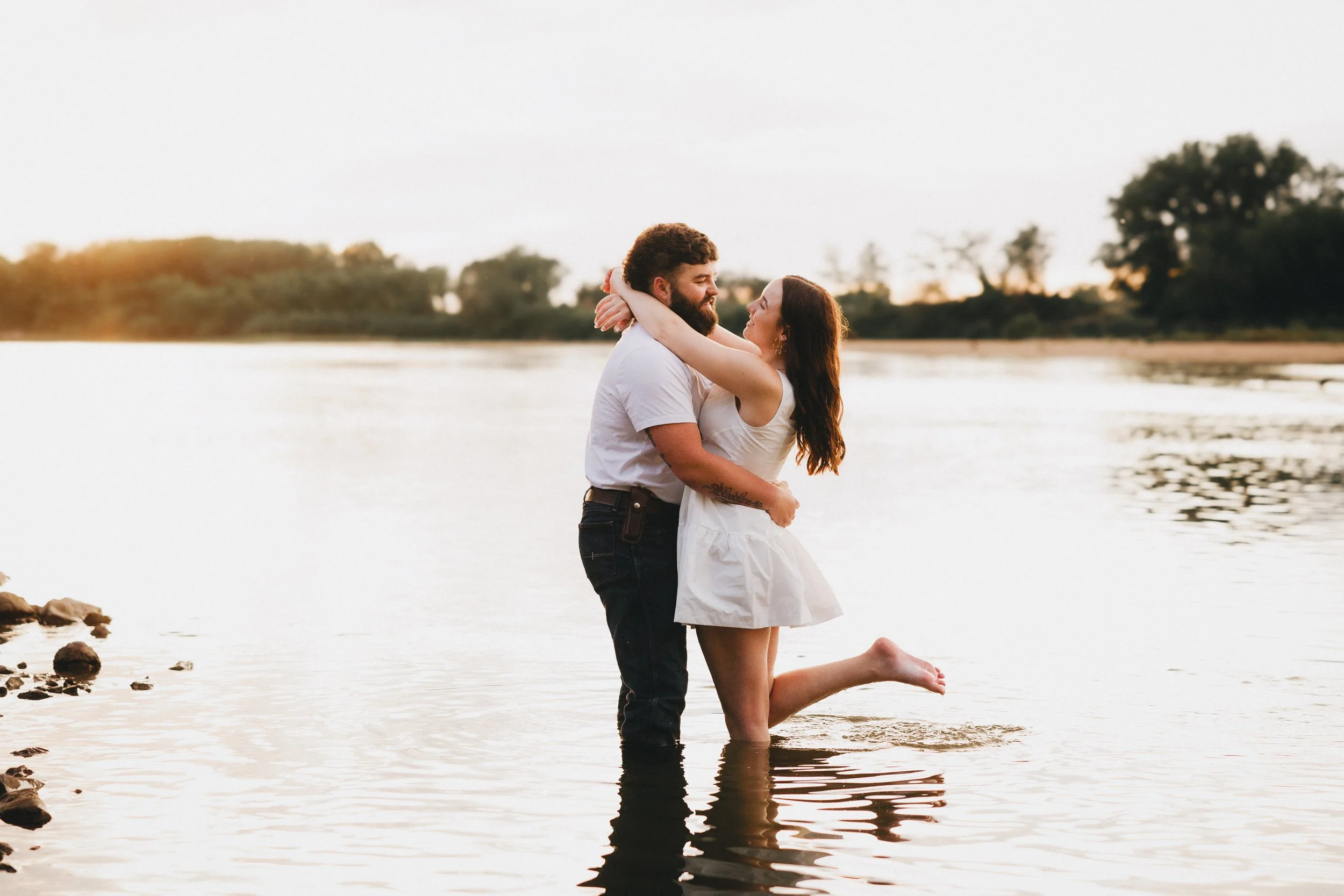 A couple embraces in shallow water at sunset, smiling and looking at each other.