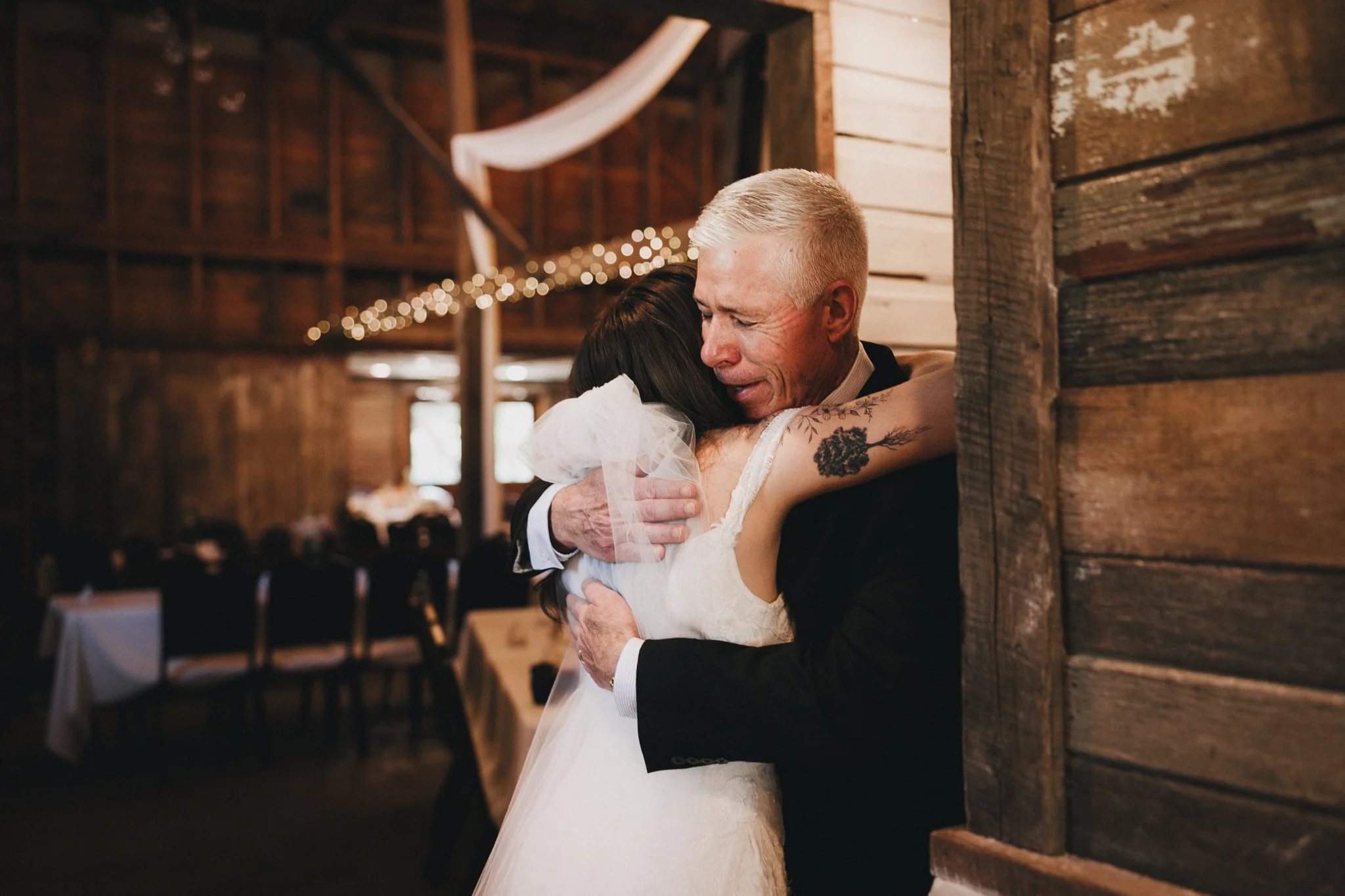 A bride and an older man, possibly her father, hugging in a rustic wedding reception space. The bride is in a white dress with a tulle veil, and the man is in a dark suit. They are sharing an emotional embrace, with warm wooden walls and string light