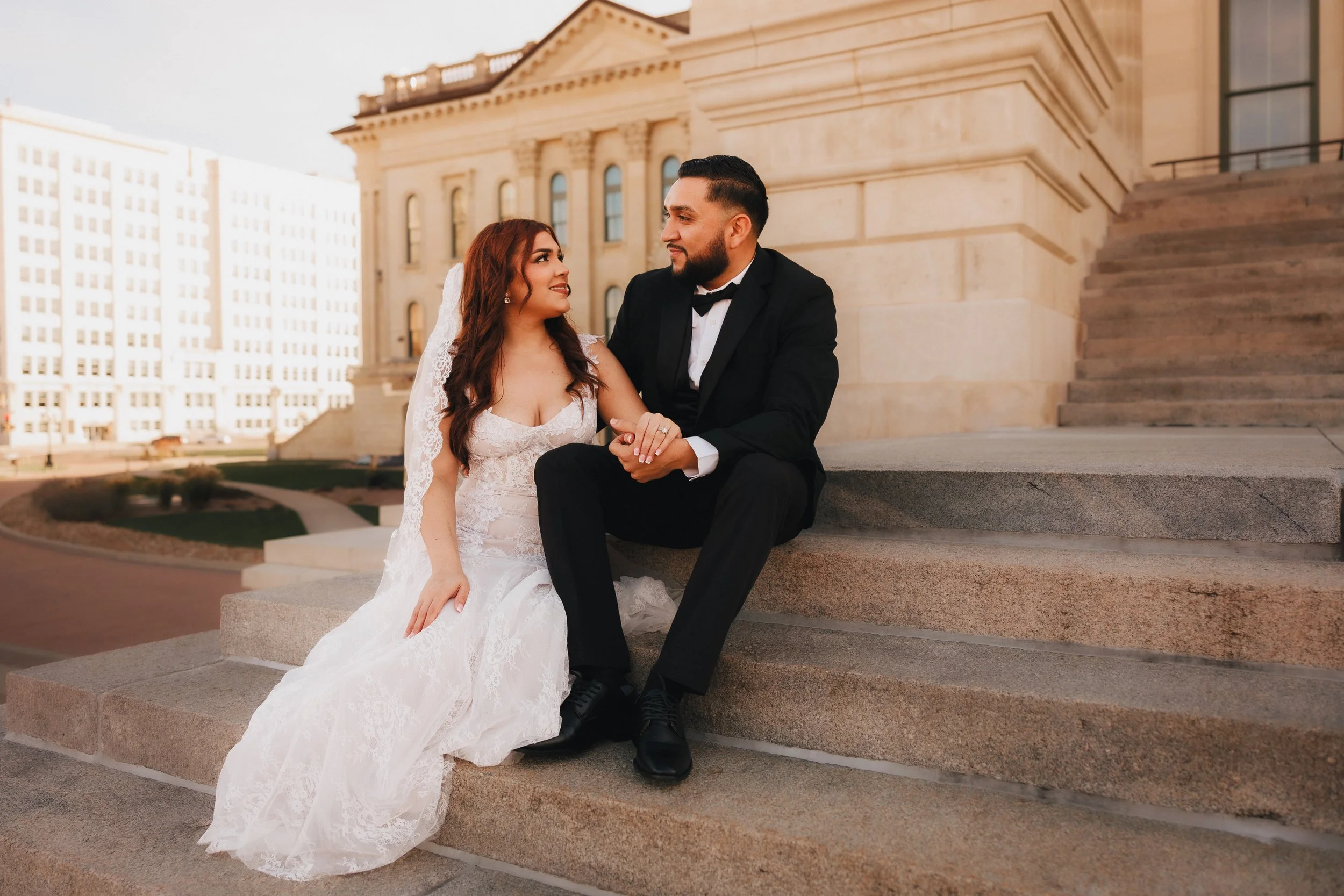 A bride and groom sitting on stairs outside Kansas State Capitol holding hands and looking at each other on their wedding day.