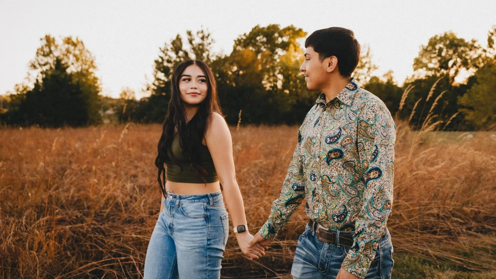 A couple walks hand in hand through a golden field at sunset in Topeka, KS, sharing a soft glance. She wears a cropped top and ripped jeans, while he sports a paisley button-up. Photo by Raw Shutter Photography.