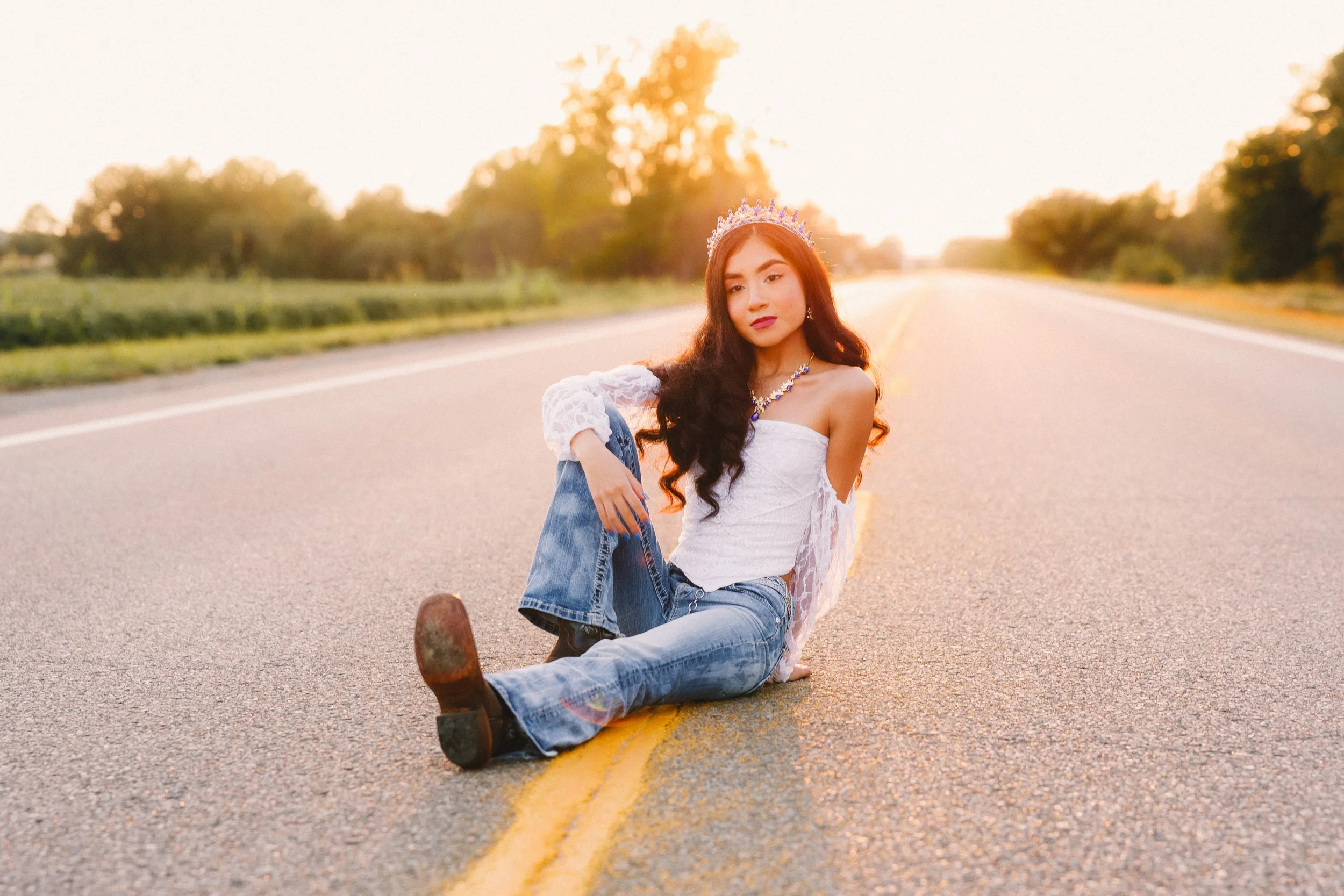 A young woman sitting on the middle of an empty road at sunset, wearing a white off-shoulder top, blue jeans, a tiara, and jewelry.