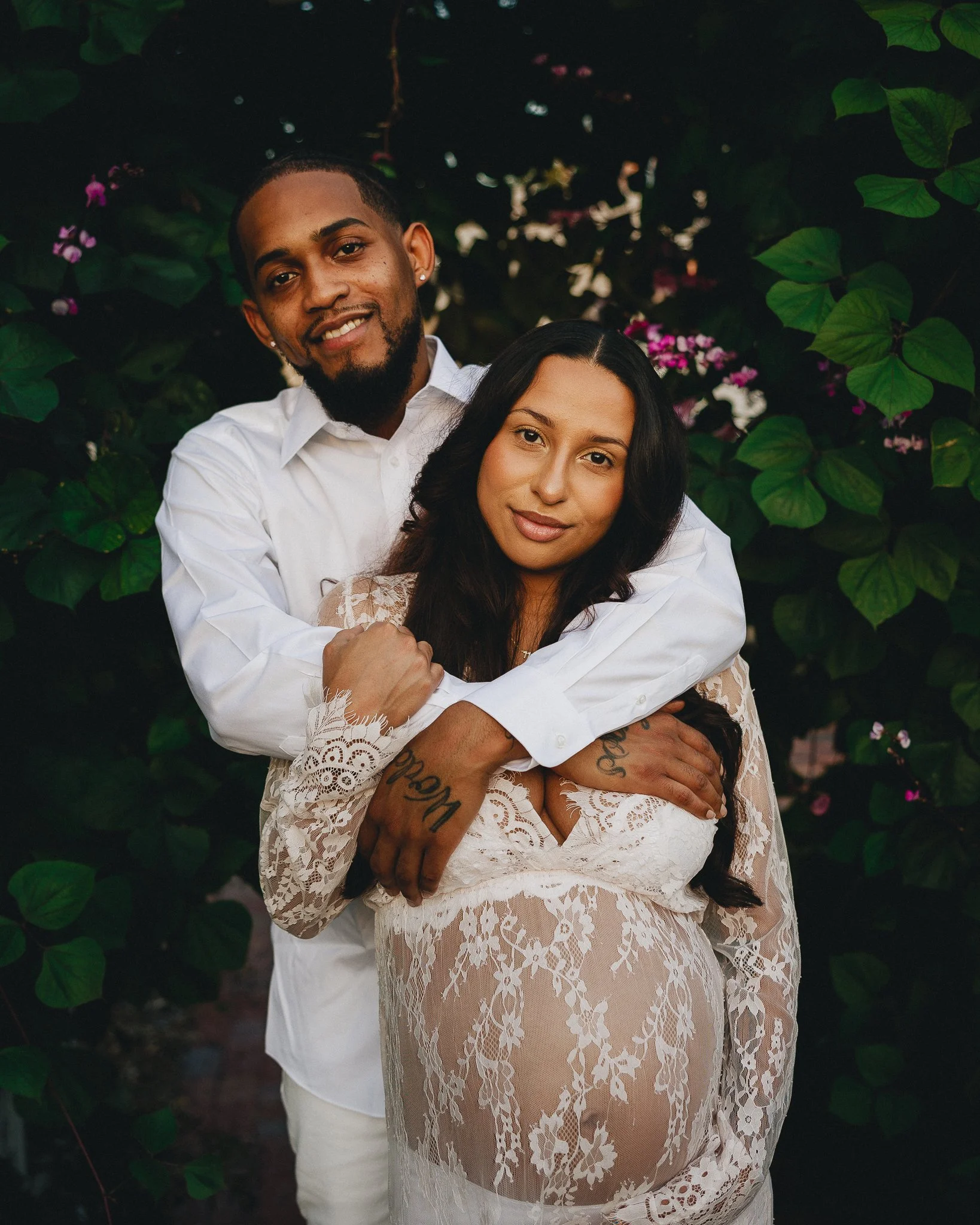 A couple is posing outdoors, with the man embracing the pregnant woman, both smiling. The woman is wearing a lace dress showing her pregnancy, and the man is wearing a white shirt. They are surrounded by green foliage and pink flowers. Shot in Topeka