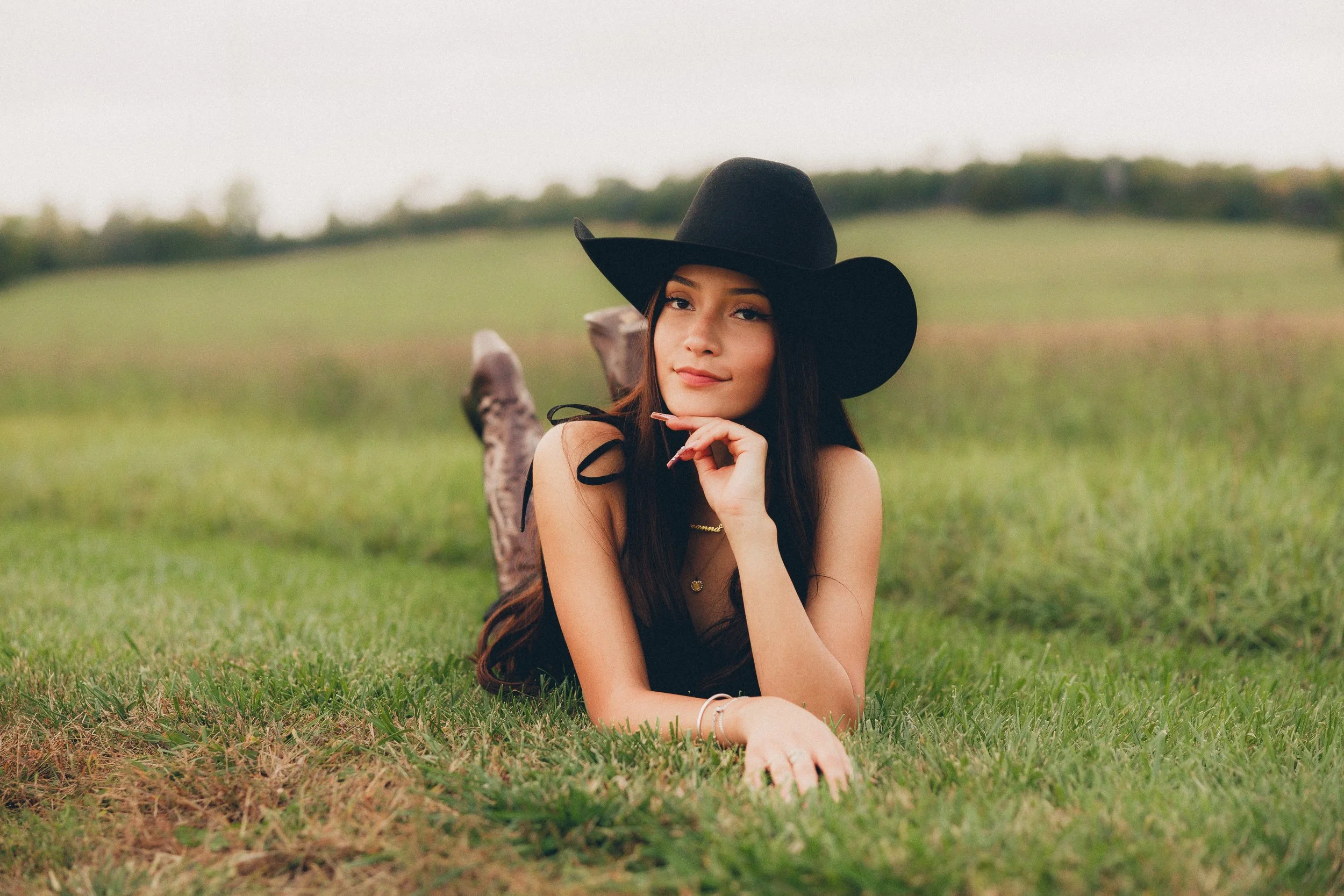 A young woman lying on green grass in an open field, wearing a large black hat and a black sleeveless dress, with her chin resting on her hand and smiling softly.