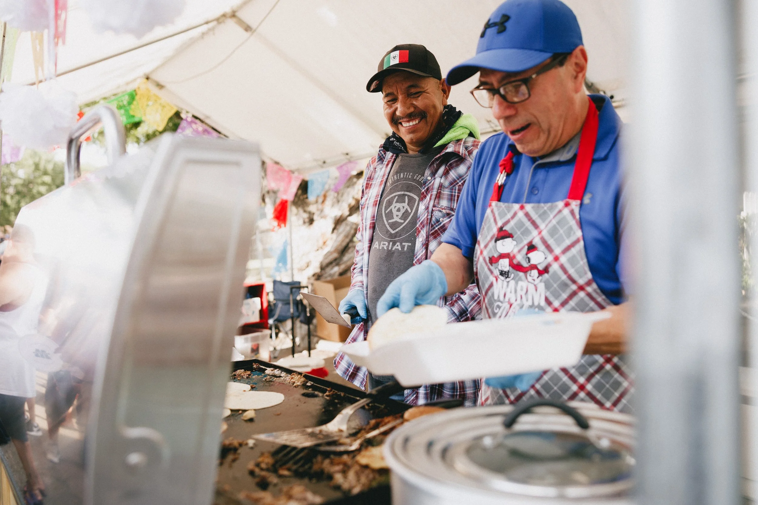 Two men serving food at an outdoor event, one wearing a blue cap and apron, the other smiling and holding a plate, under a tent. Shot at Fiesta Topeka.