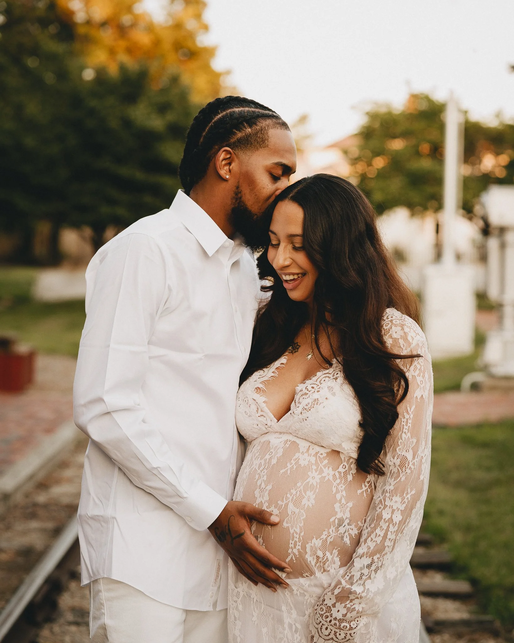A couple embracing outdoors, with the man kissing the woman's forehead as she smiles, and the woman's pregnant belly visible in a lace dress. Shot in Topeka, Kansas. 