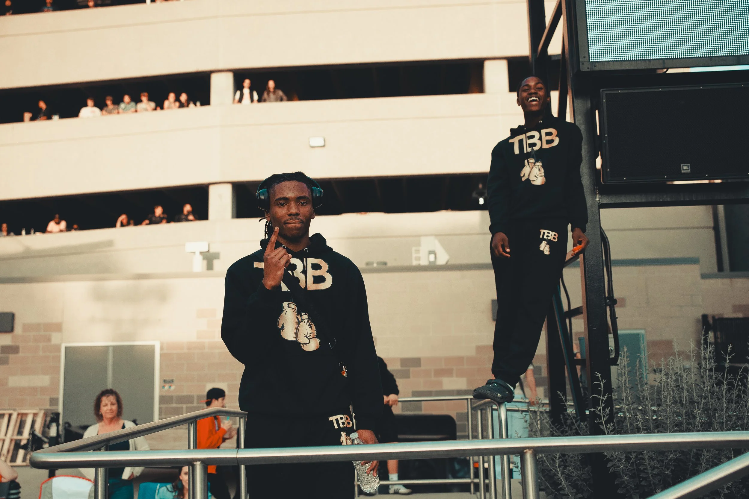 Two young men in black hoodies with "TBB" logo standing outdoors, one holding a water bottle and the other on a staircase. People are visible in the background on multiple levels of a building. Shot at Evergy Plaza in Topeka, Kansas. 