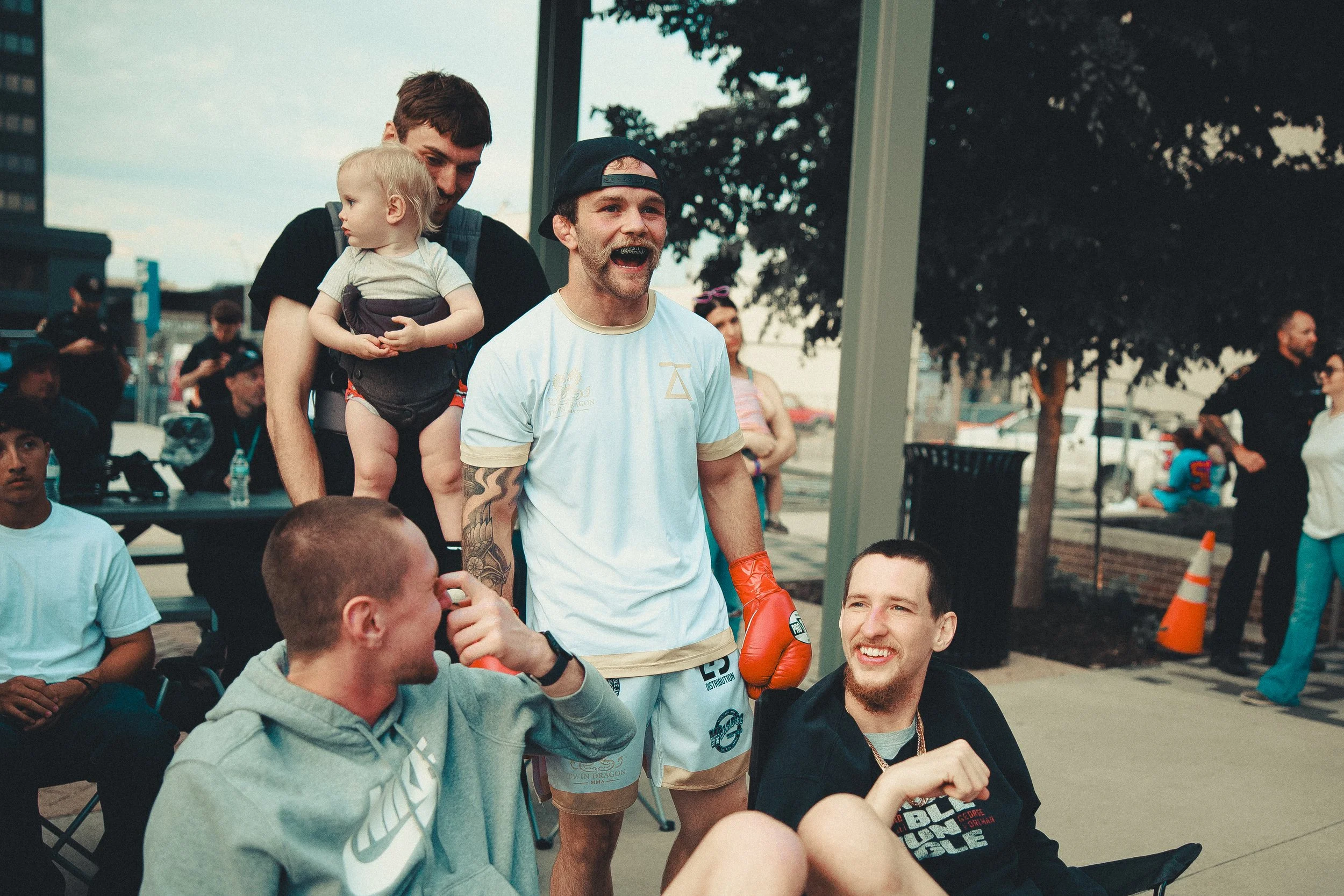 A group of men and women at a public outdoor event, some sitting and some standing, with a man in boxing gloves smiling and talking to others. Shot at Evergy Plaza in Topeka, Kansas. 