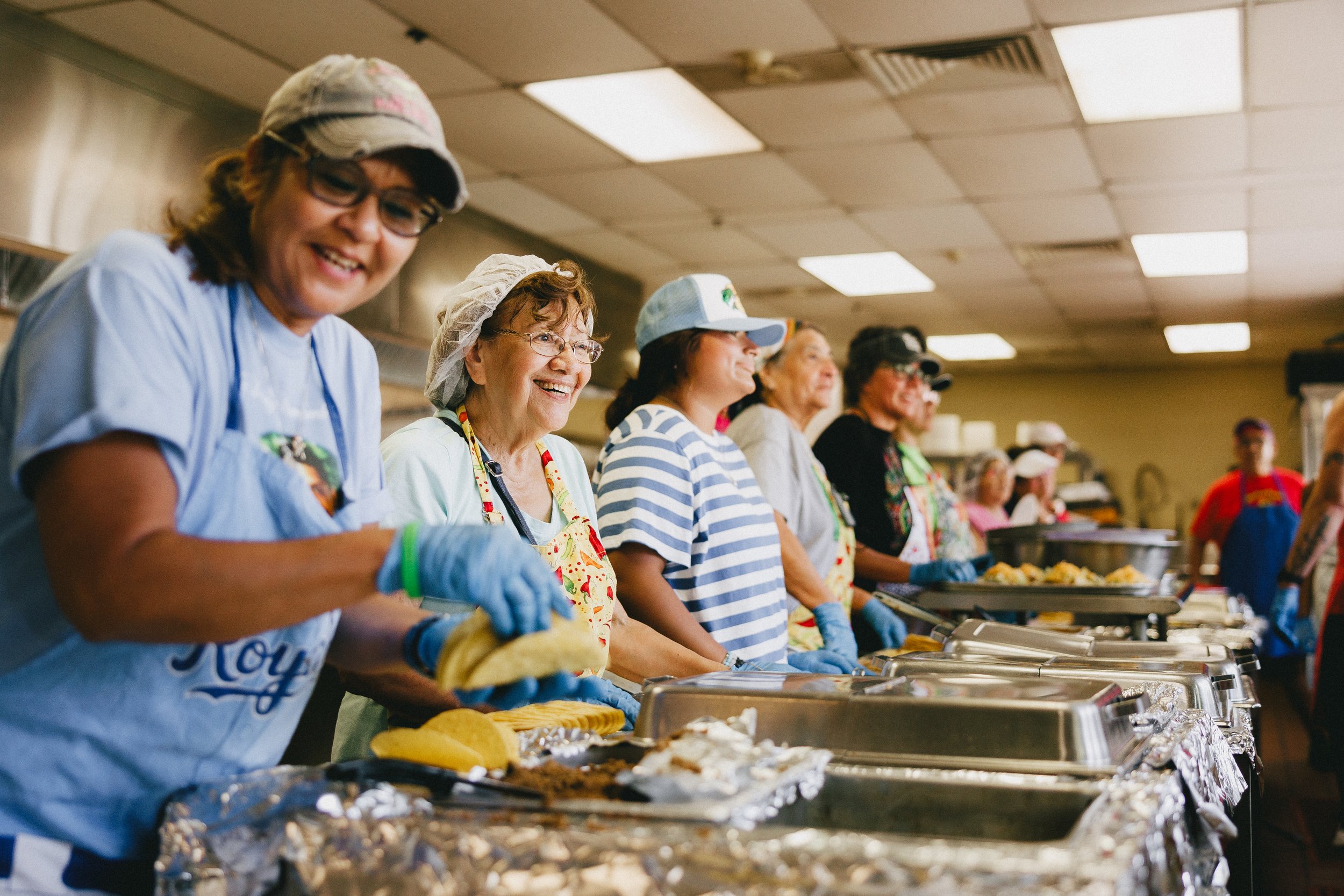 A group of smiling women in aprons and gloves serving food at a buffet line in a community kitchen or event hall. Shot at Fiesta Topeka.
