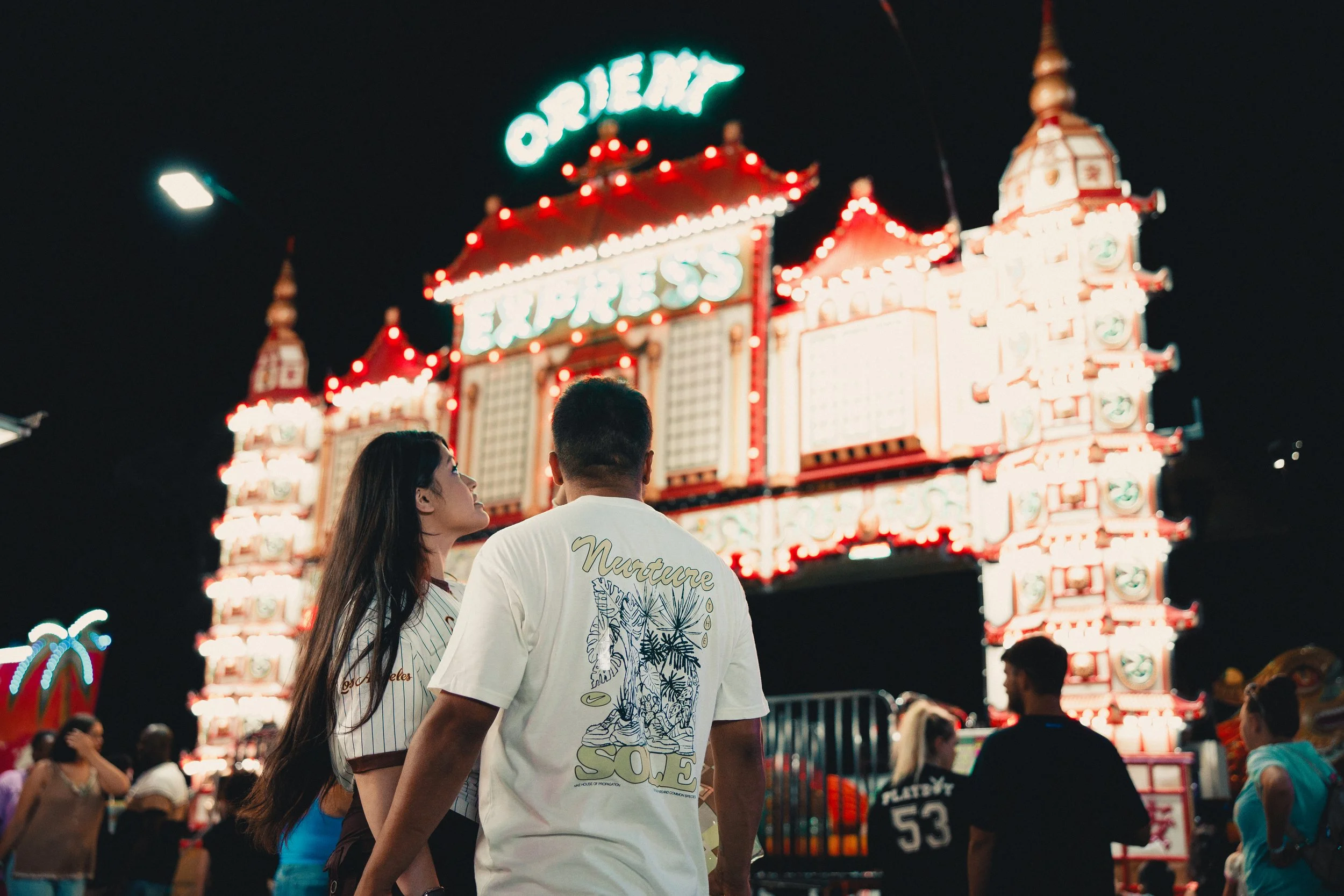 People at a night fairground, with a brightly lit amusement ride or game booth in the background. Shot at Fiesta Topeka.