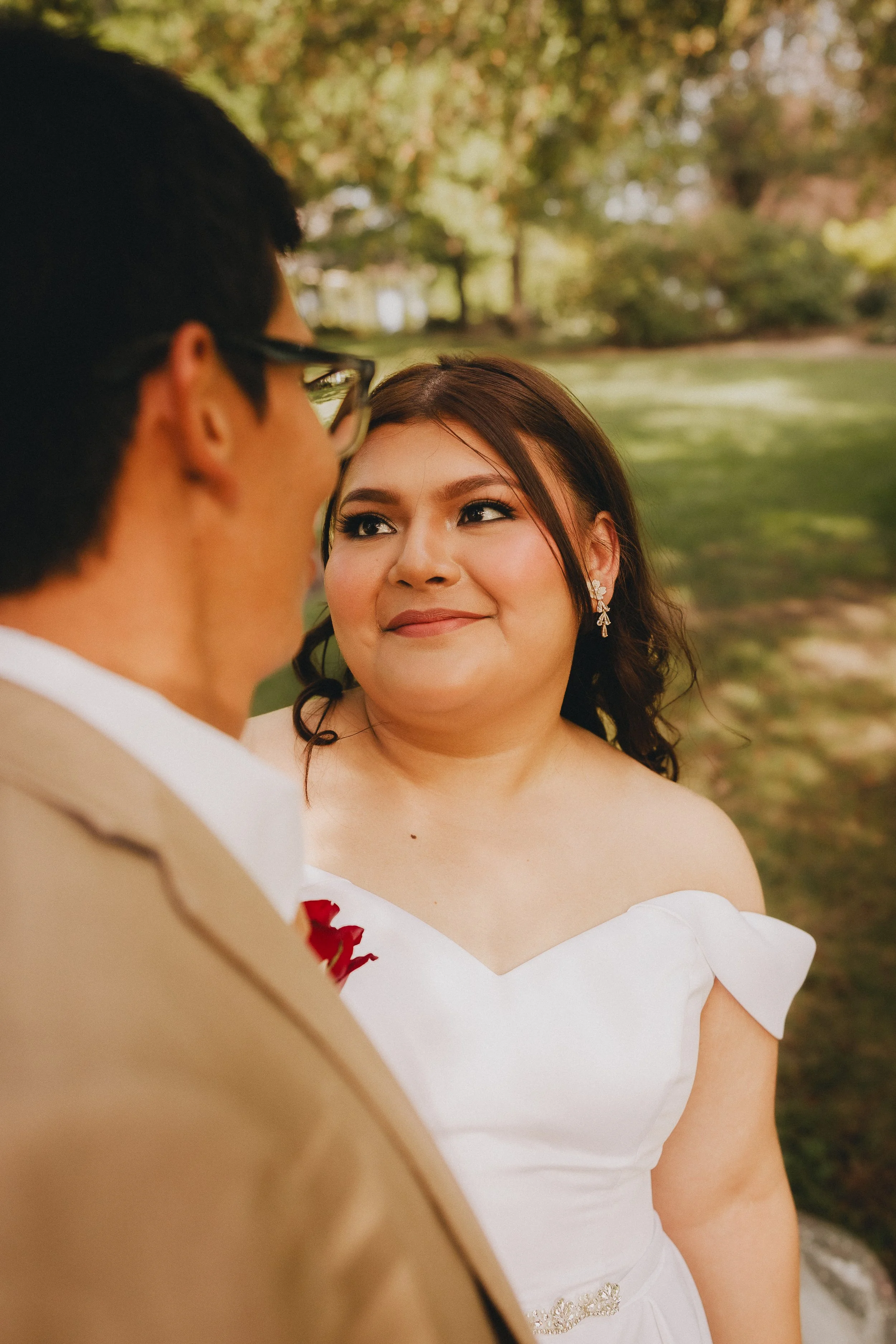 A woman in a white wedding dress with dark hair and earrings looks at a man in a tan suit with glasses during an outdoor wedding ceremony in a park with green trees and grass.