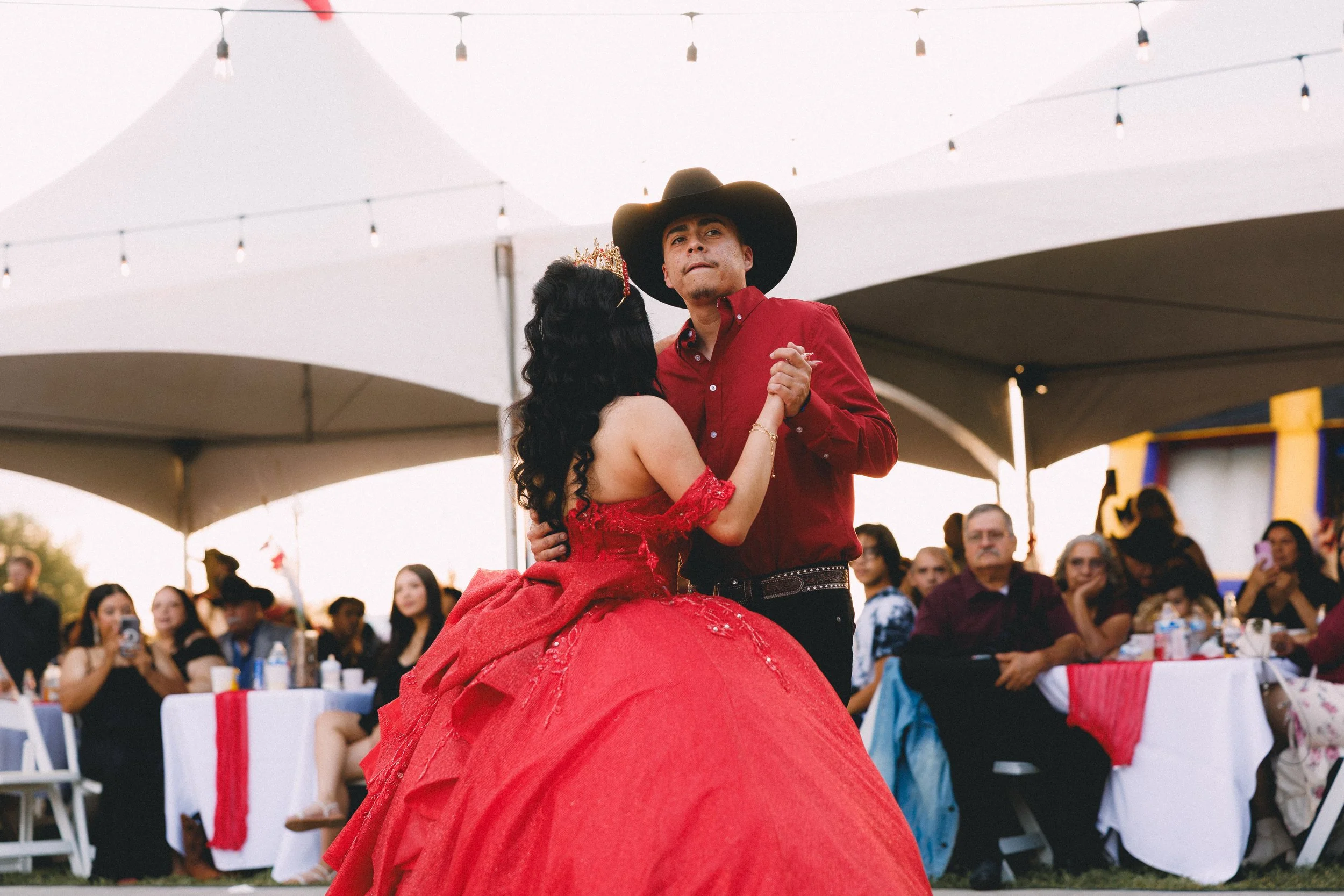 A young woman in a red ball gown and a young man in a red shirt with a cowboy hat are dancing together at an outdoor event, surrounded by seated guests and under large tents.